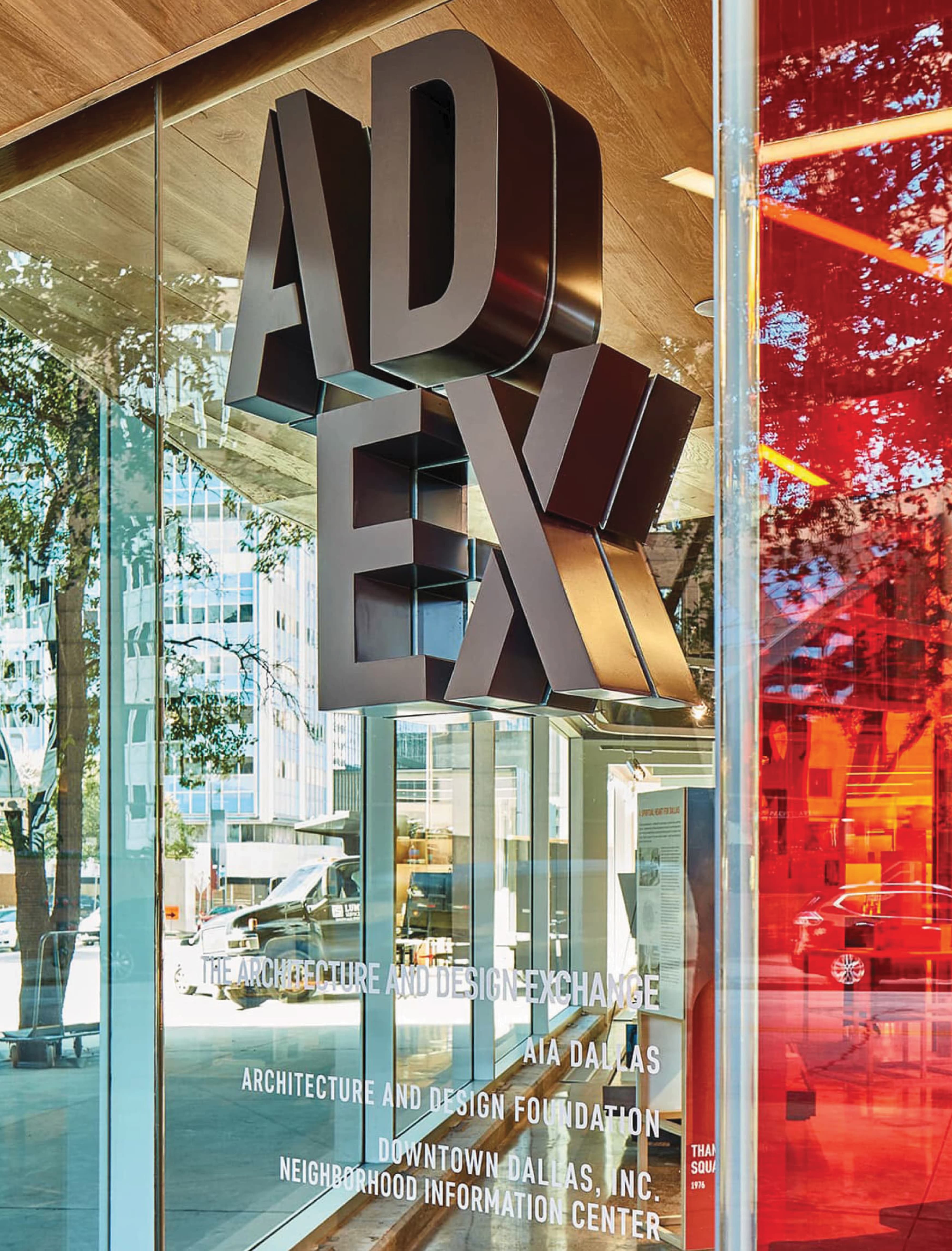 RSM Design entry identity signage and window graphics for the Architecture and Design Exchange in downtown Dallas, featuring a close-up of large-scale suspended dimensional dark bronze ADEX letter forms mounted beneath a wood canopy with downtown Dallas reflected in the glass facade, alongside a full tenant directory window graphic listing AIA Dallas, Architecture and Design Foundation, Downtown Dallas Inc., and Neighborhood Information Center, demonstrating how environmental graphic design, visual identity, wayfinding and signage design, and branded environments create a layered, informative, and architecturally integrated entry experience for cultural, civic, and urban streetscape markets.