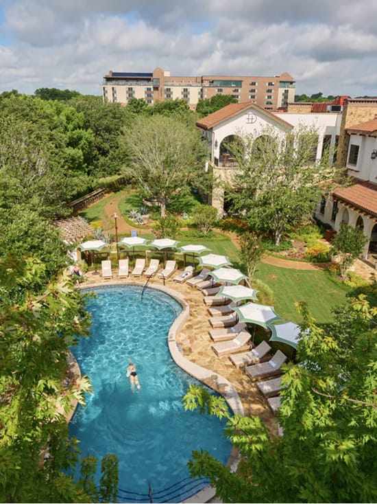 Aerial view of Hotel Drover's outdoor pool and sundeck in Fort Worth Stockyards, Texas, featuring a freeform swimming pool, lounge chairs with green-striped umbrellas, stone terrace, and lush landscaped grounds surrounding the hotel's Spanish Colonial-style architecture