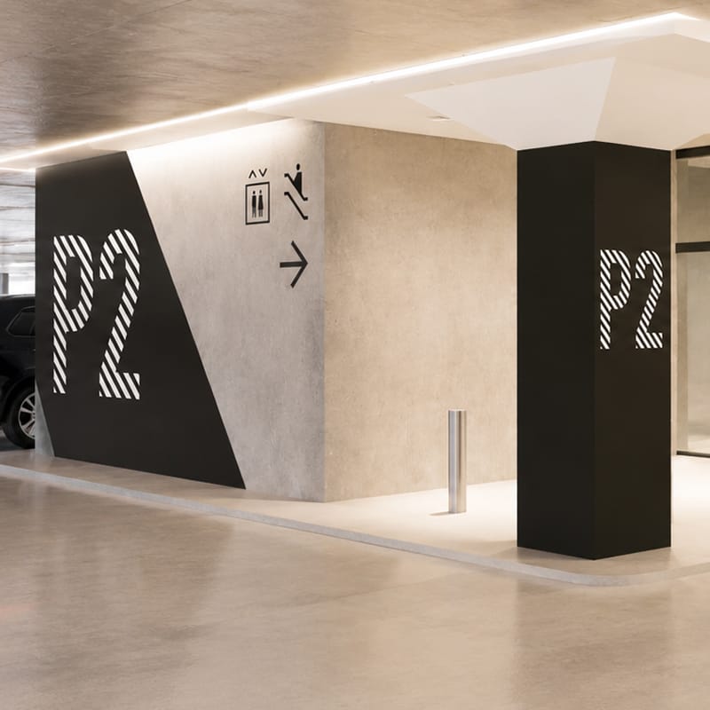 Parking garage level identification signage with bold striped "P2" graphics on a black angled wall and column, elevator and escalator pictograms nearby — parking wayfinding design by RSM Design