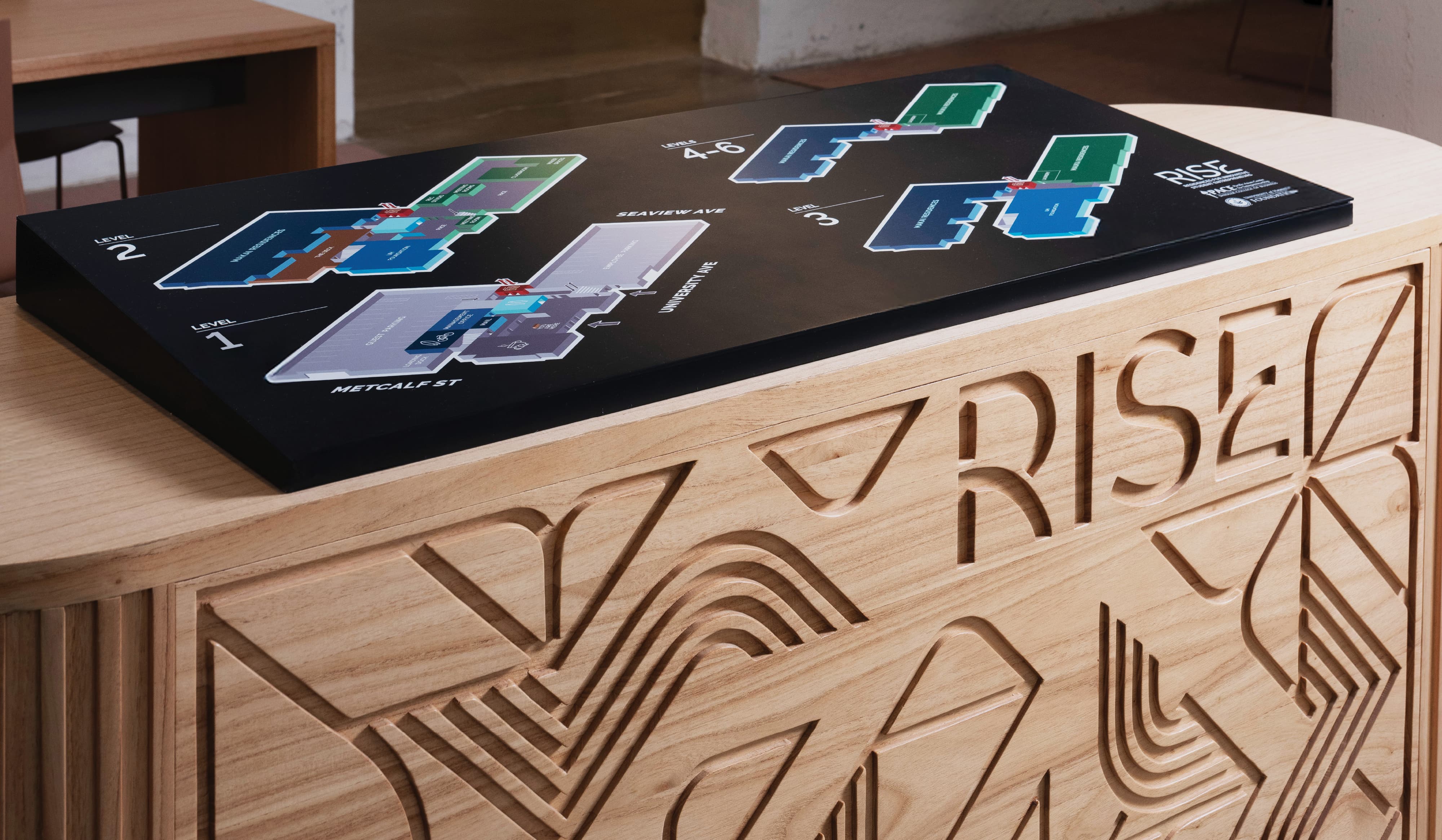 Wooden reception desk carved with the 'RISE' logo and a mountain-and-wave pattern, topped by a black floor plan directory showing Levels 1 through 6 with color-coded building sections along Metcalf Street, Seaview Avenue, and University Avenue.