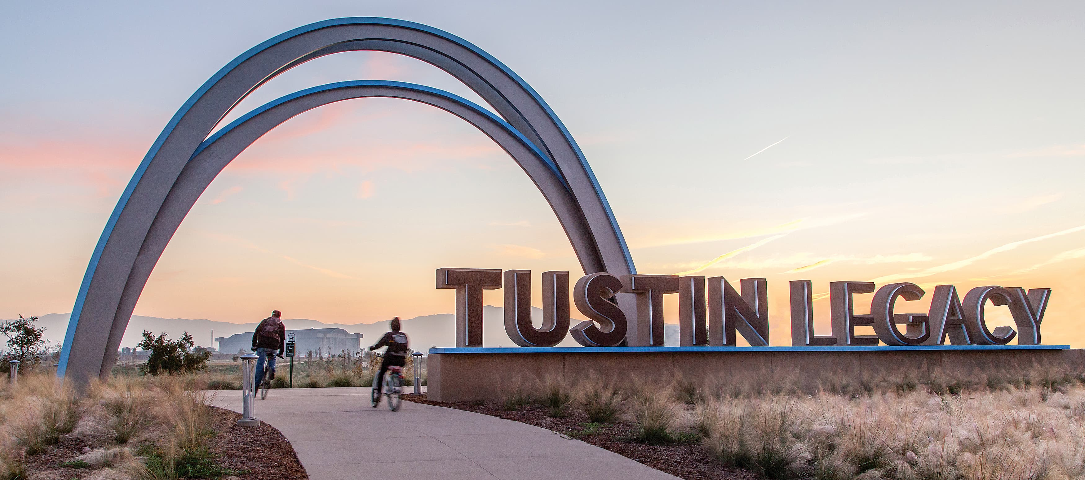 Two people riding bikes under a large arch that says Tustin Legacy.