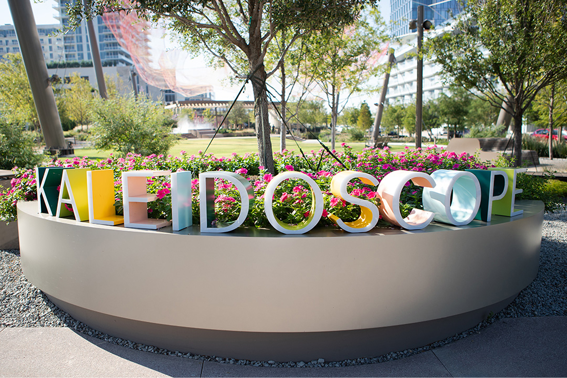 Colorful multicolored "Kaleidoscope" dimensional letter sculpture in circular planter with pink flowers at Kaleidoscope Park public plaza, Frisco, Texas