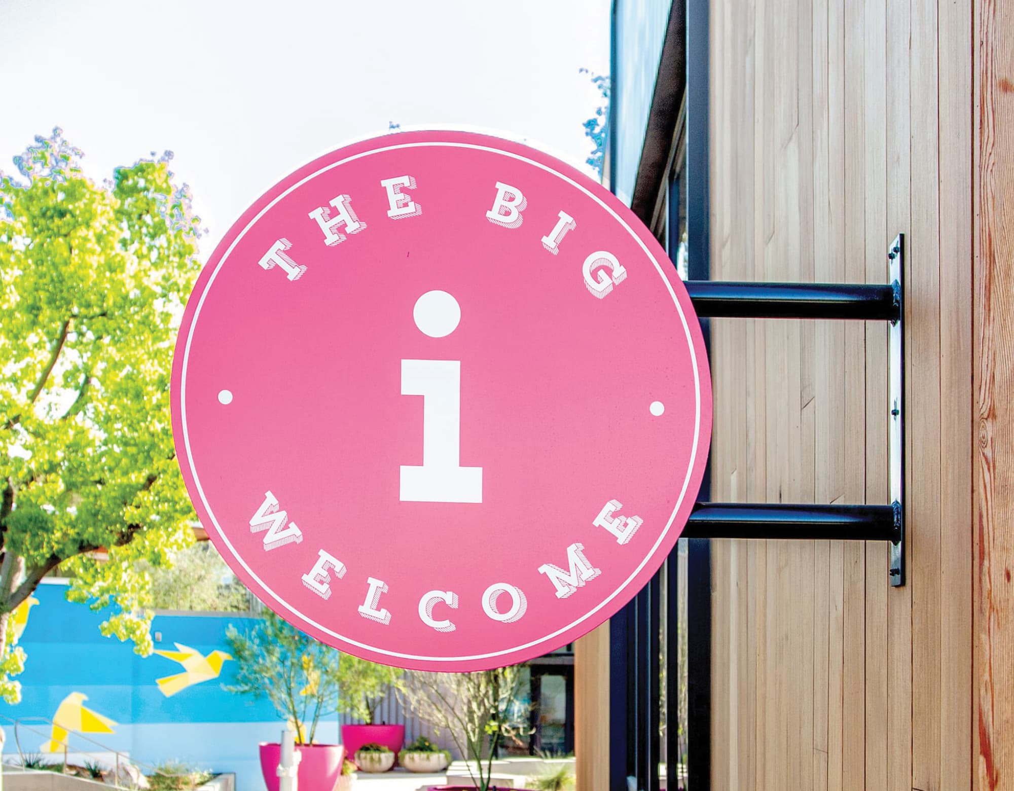 Round pink blade sign reading "The Big Welcome" with an information icon mounted on a wood-clad exterior wall at a residential community, highlighting placemaking, wayfinding, and branded environment design by RSM Design.