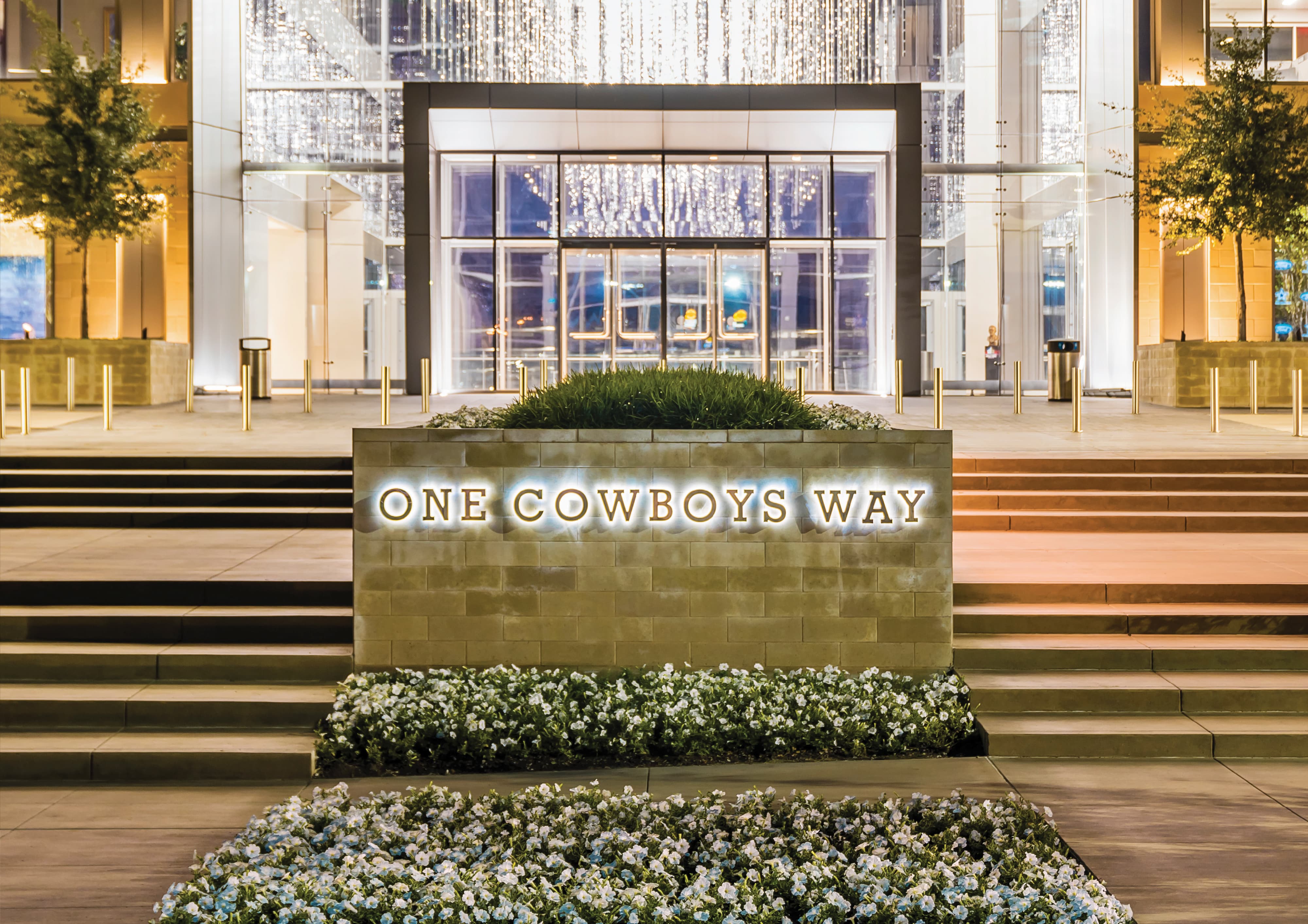Illuminated ground-level monument sign reading 'One Cowboys Way' set within a landscaped planter at the entrance to The Star in Frisco, Texas — an example of RSM Design's placemaking and environmental graphic design for a mixed-use sports and entertainment destination.