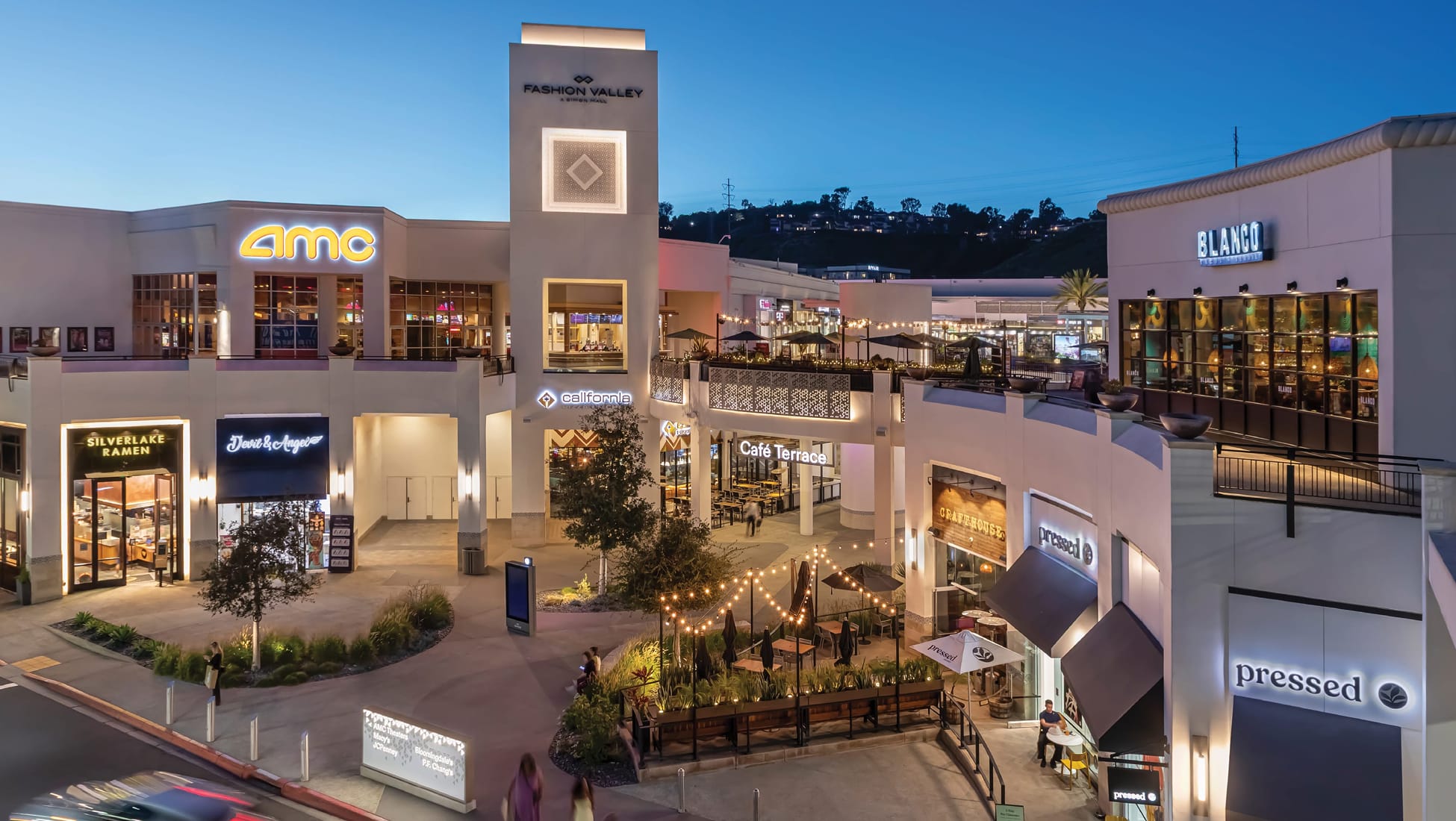 Aerial dusk view of Fashion Valley Mall's open-air dining and entertainment district in San Diego, featuring string-lit outdoor seating, the central Fashion Valley tower, and tenants including AMC, Silverlake Ramen, Pressed, and Blanco against a deep blue evening sky.