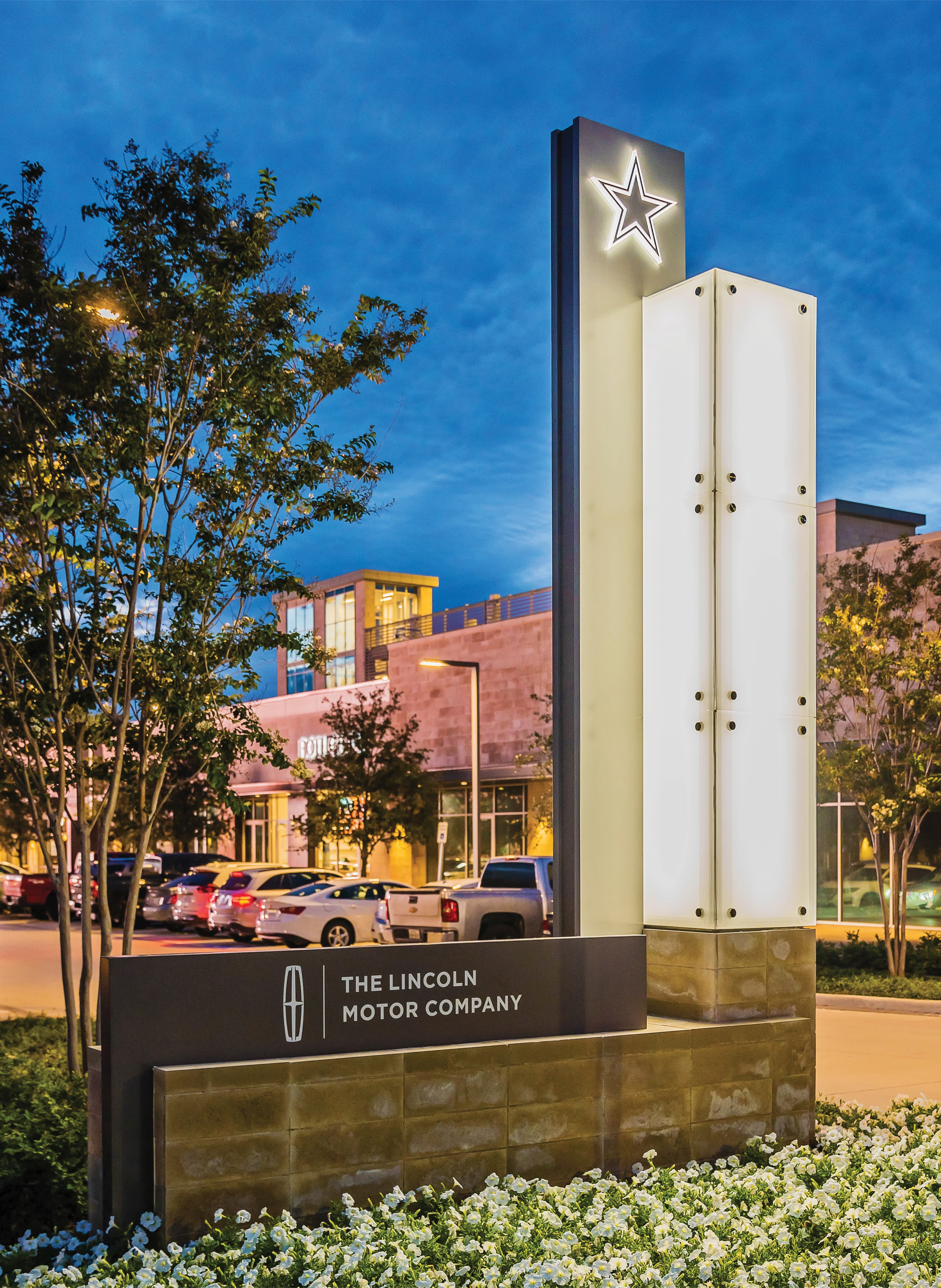 A dramatic dusk view of an illuminated entry monument sign at The Star in Frisco featuring a backlit translucent tower with a glowing Dallas Cowboys star topper, and a stone base panel identifying The Lincoln Motor Company as a sponsor, set among white flowering landscaping and mature trees with the retail district warmly lit in the background — illustrating RSM Design's identity signage, master sign programs, and placemaking services applied within the sports and entertainment and mixed-use markets