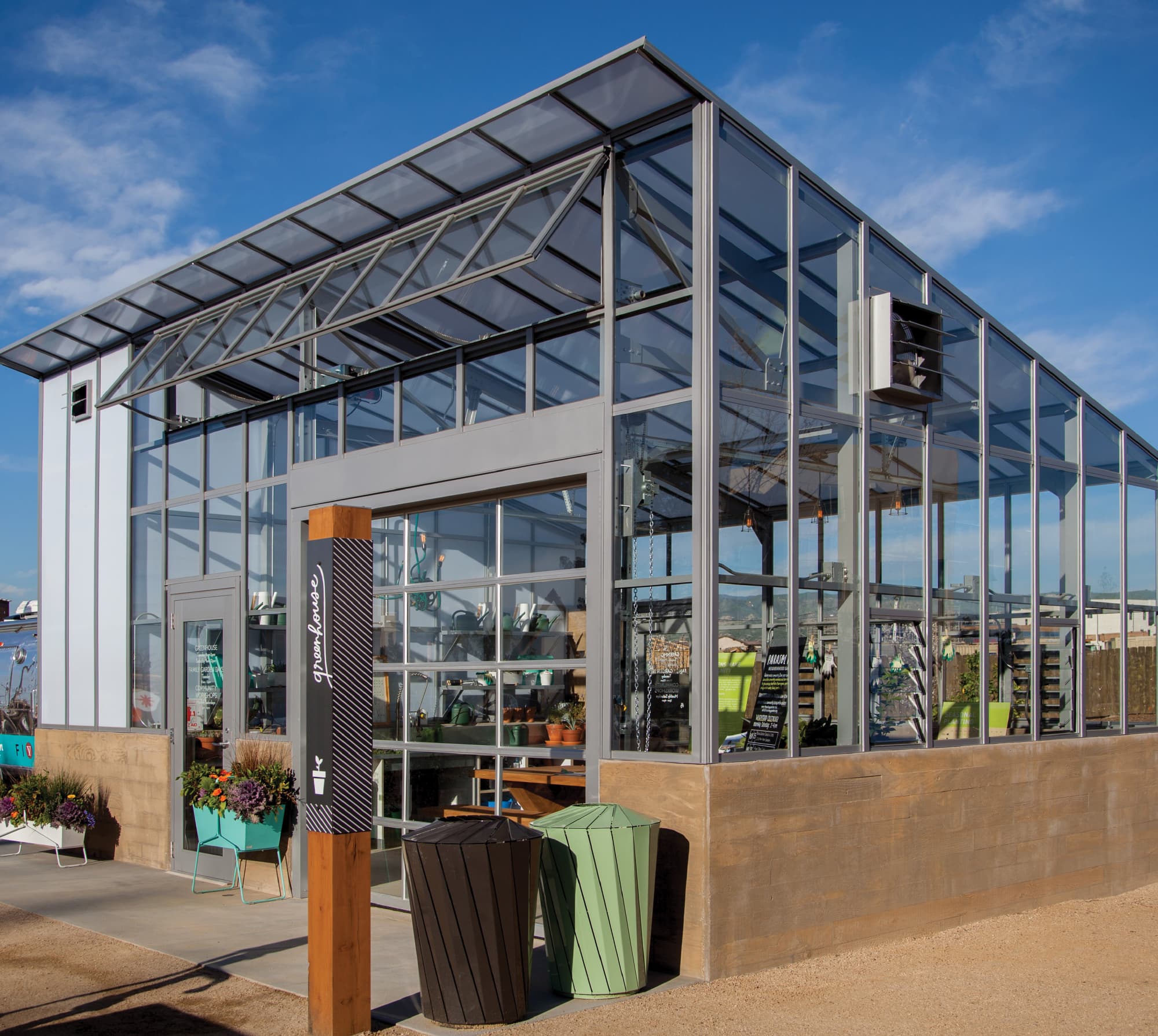 RSM Design amenity identification signage and placemaking for FivePoint at Great Park Neighborhoods in Irvine, California, featuring a branded black wood post sign with vertical script "greenhouse" lettering and diagonal stripe pattern at the entry of a glass and steel community greenhouse structure, with colorful planters and coordinating waste receptacles in the foreground against a bright blue sky, demonstrating how environmental graphic design, specialty graphics, wayfinding and signage design, and placemaking extend a cohesive visual identity across every amenity touchpoint to create a distinctly human-centered branded environment for residential community and park markets.