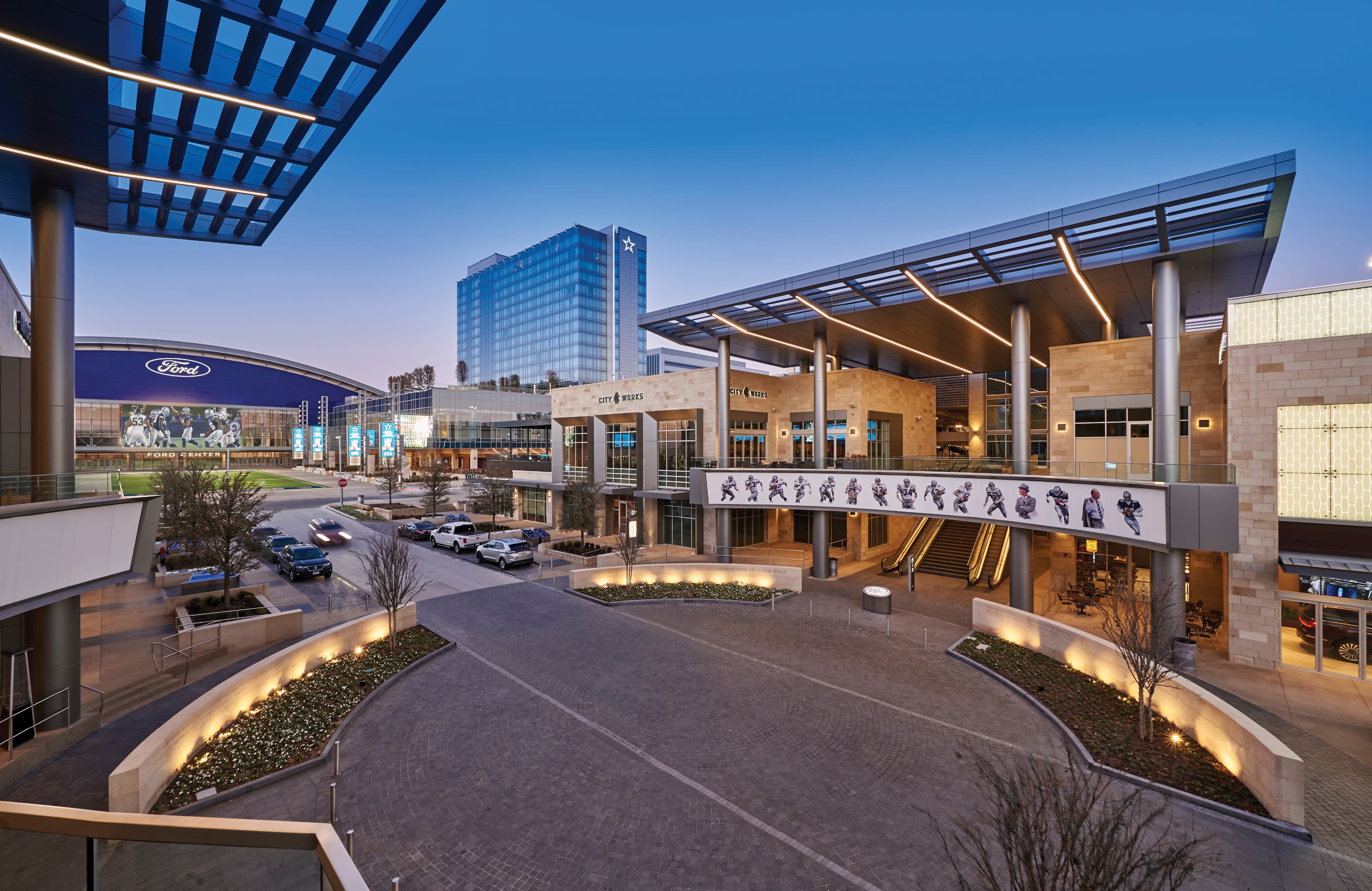 Aerial dusk view of The Star in Frisco, Texas — a mixed-use sports and entertainment destination featuring the Ford Center at The Star, large-scale experiential graphics of Dallas Cowboys players along a pedestrian walkway, City Works restaurant signage, and the Cowboys star atop a corporate office tower — showcasing RSM Design's integrated approach to environmental graphic design, placemaking, and branded environments.