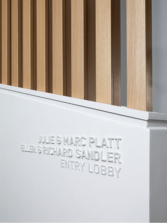 Close-up architectural detail of the Julie and Marc Platt and Ellen and Richard Sandler Entry Lobby donor recognition signage featuring tonal white embossed dimensional lettering on a white wall surface, with a natural oak vertical wood slat screen above, showcasing refined interior wayfinding and named space design