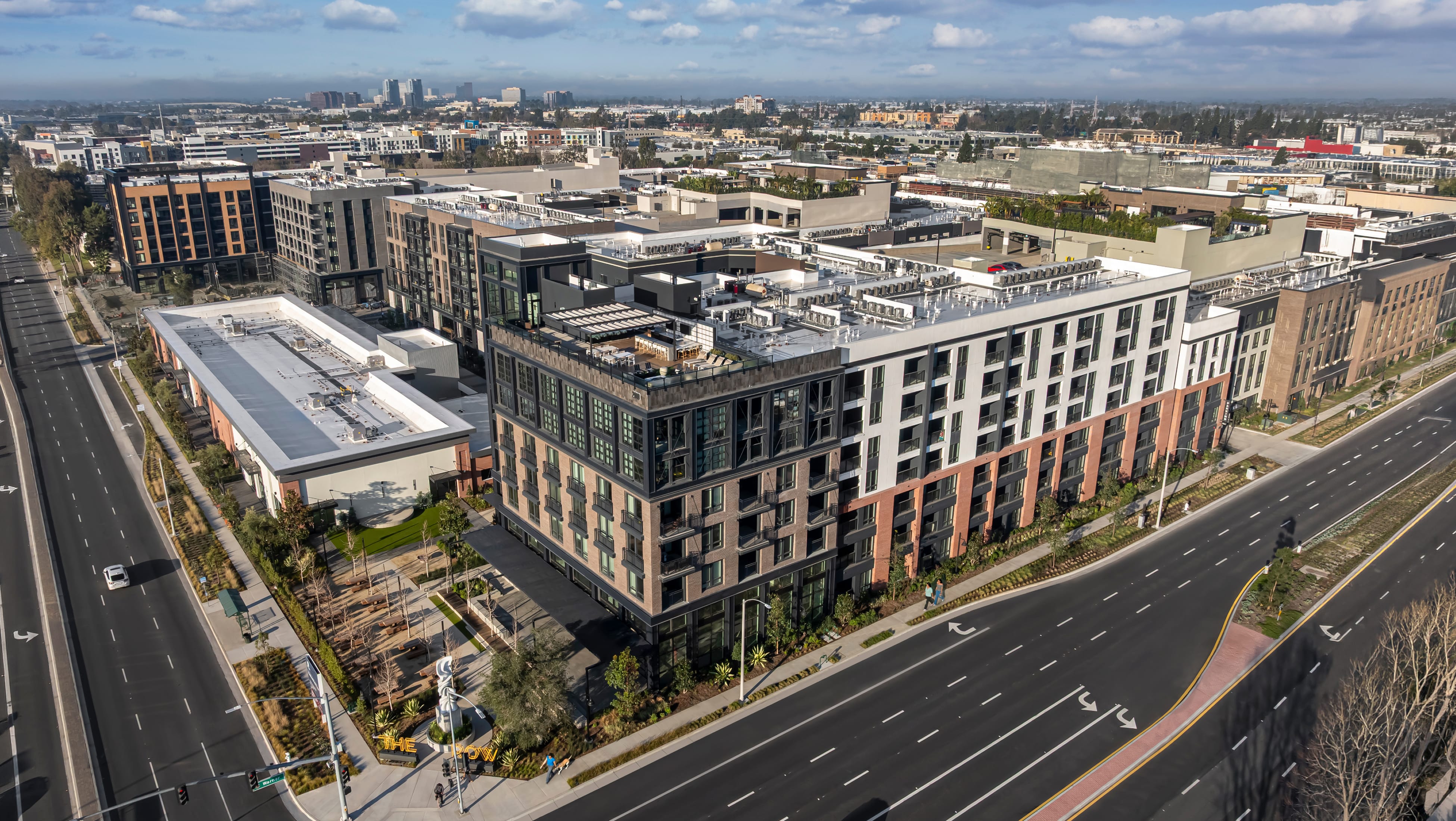 Aerial view of The Row at Red Hill mixed-use residential development, showing a brick and dark-metal apartment building with a rooftop terrace, ground-level plaza, and illuminated "THE ROW" monument sign along Red Hill Avenue – placemaking and environmental graphic design by RSM Design.