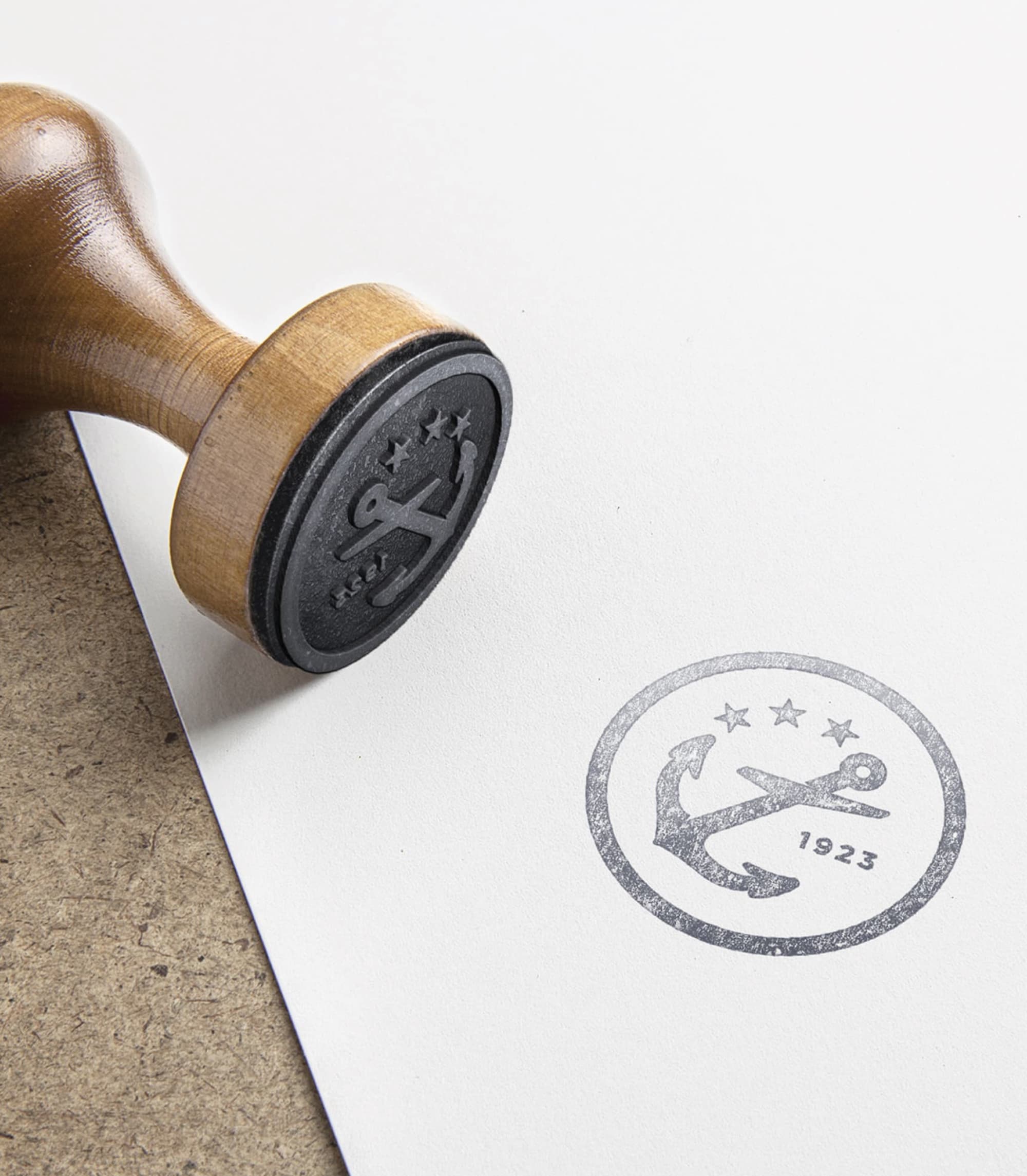 Wooden-handled rubber stamp beside its printed impression on paper, showing a circular anchor emblem with three stars and '1923' text.