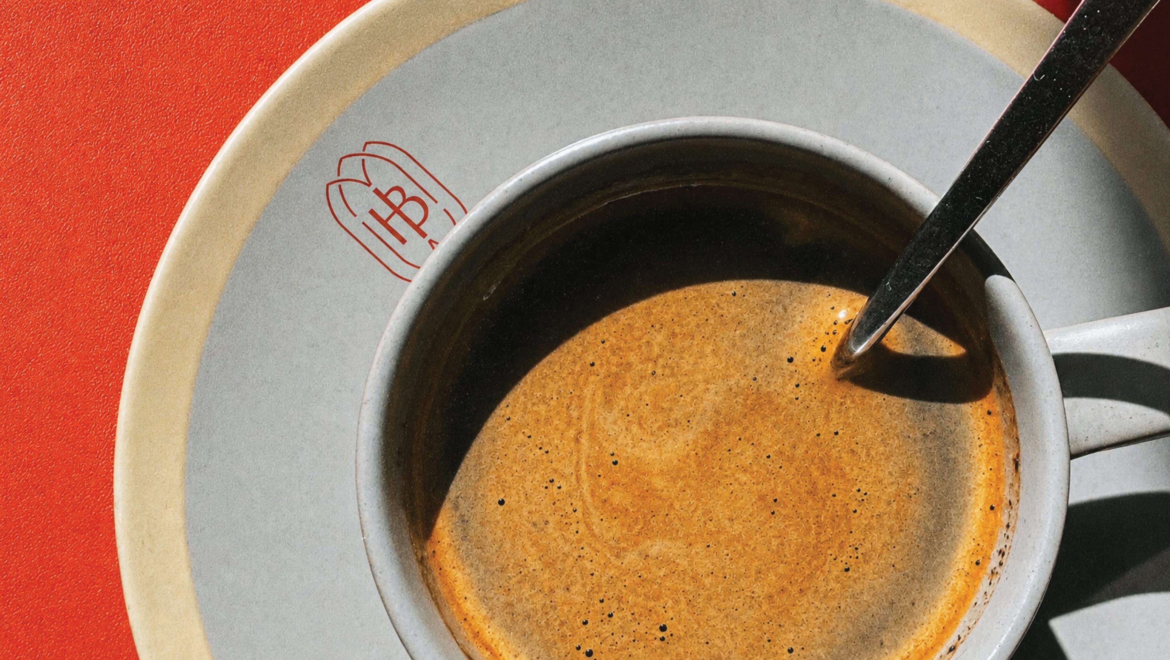 Close-up overhead shot of espresso in a branded cup with heart-shaped "HB" logo mark on saucer, red tabletop — brand identity design by RSM Design