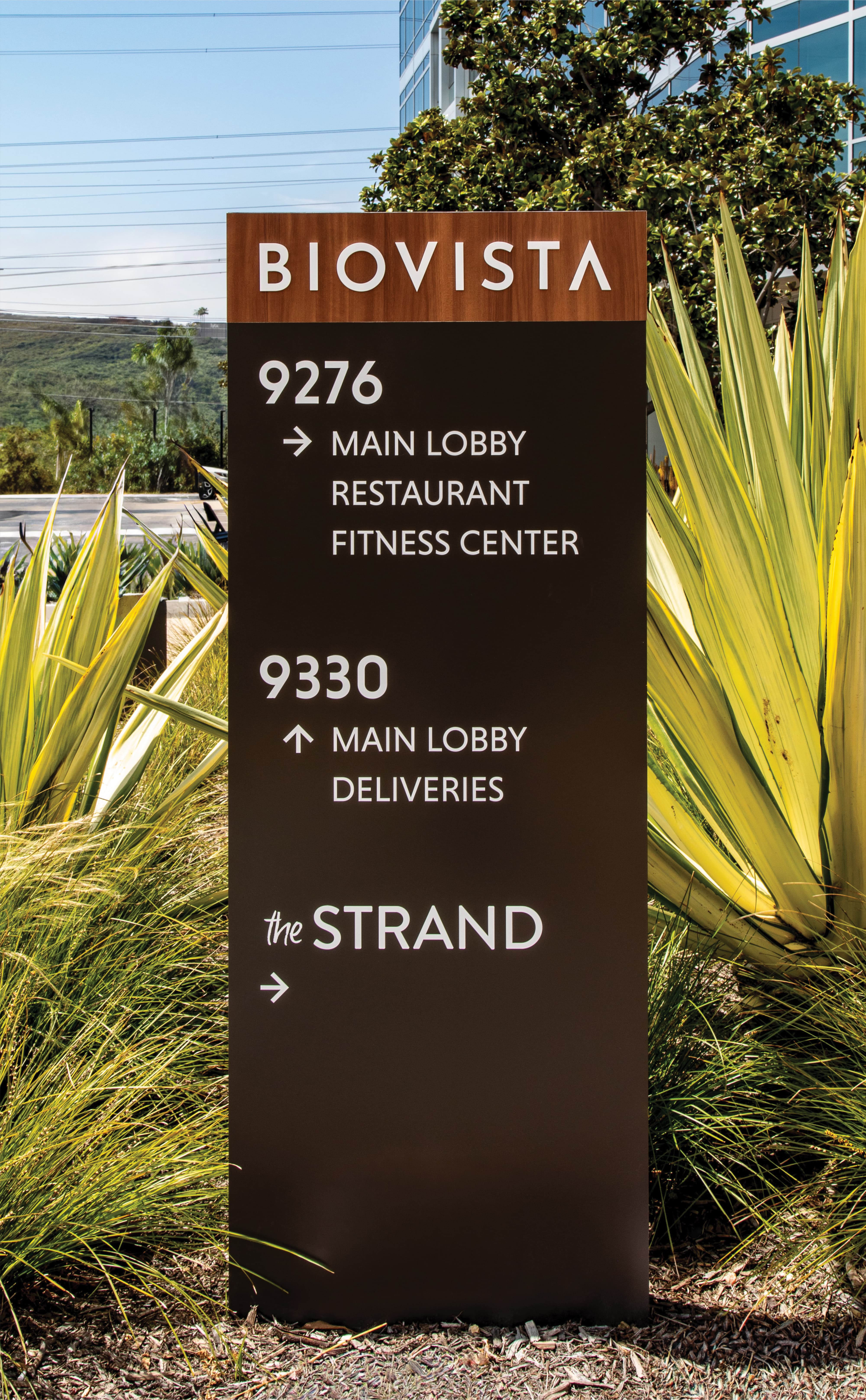 Tall wayfinding pylon sign reading 'Biovista' at the top with a wood-grain header, listing directions below to '9276: Main Lobby, Restaurant, Fitness Center,' '9330: Main Lobby, Deliveries,' and 'the Strand,' set among desert grasses and yucca plants.