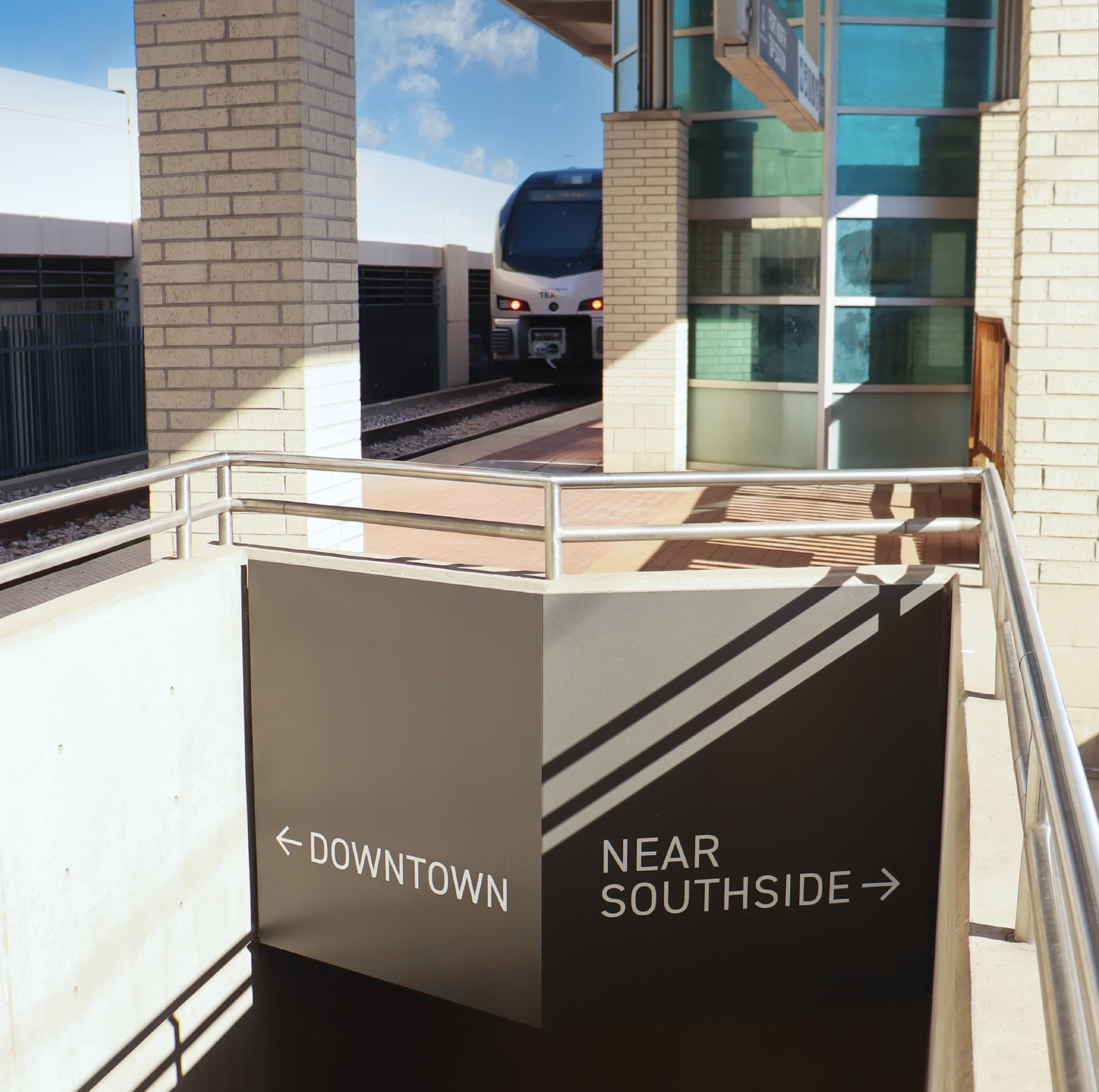 Directional wayfinding signage on a stairwell wall guiding passengers to Downtown and Near Southside at T&P Station with a commuter train visible on the platform, showcasing environmental graphic design and signage for a transit development by RSM Design.
