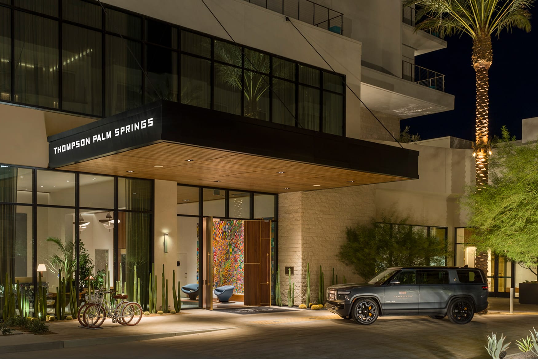Nighttime porte-cochère entrance of Thompson Palm Springs hotel, with a wood-clad canopy, illuminated hotel signage, columnar cacti, and a colorful mosaic artwork visible through the open lobby doors.