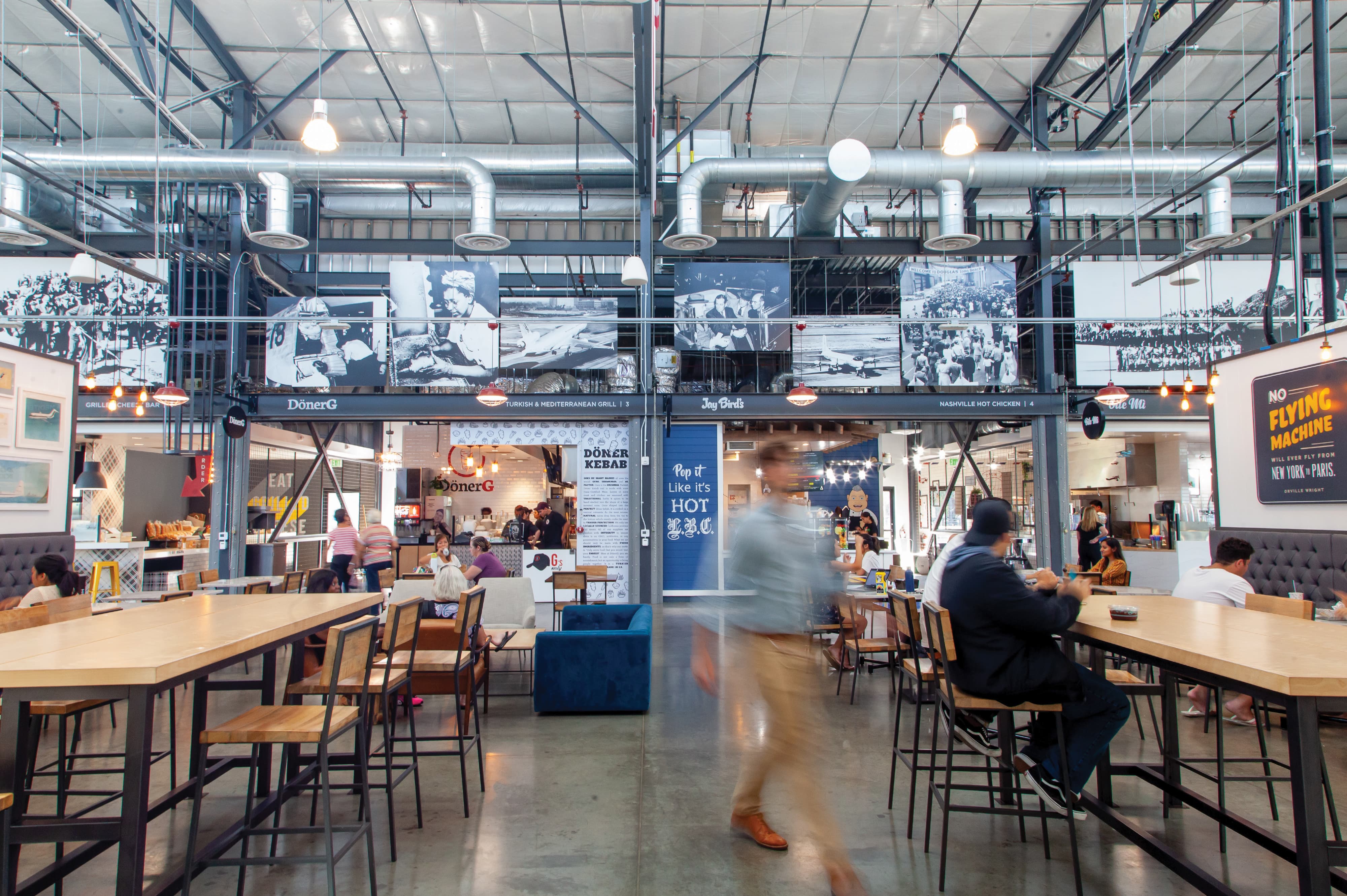 Industrial food hall interior with communal wood tables, historic black-and-white aviation photographs mounted overhead, food stalls including DönerG and Jay Bird's Nashville Hot Chicken, and a 'No Flying Machine' poster on the right, as diners sit and a blurred man walks through.