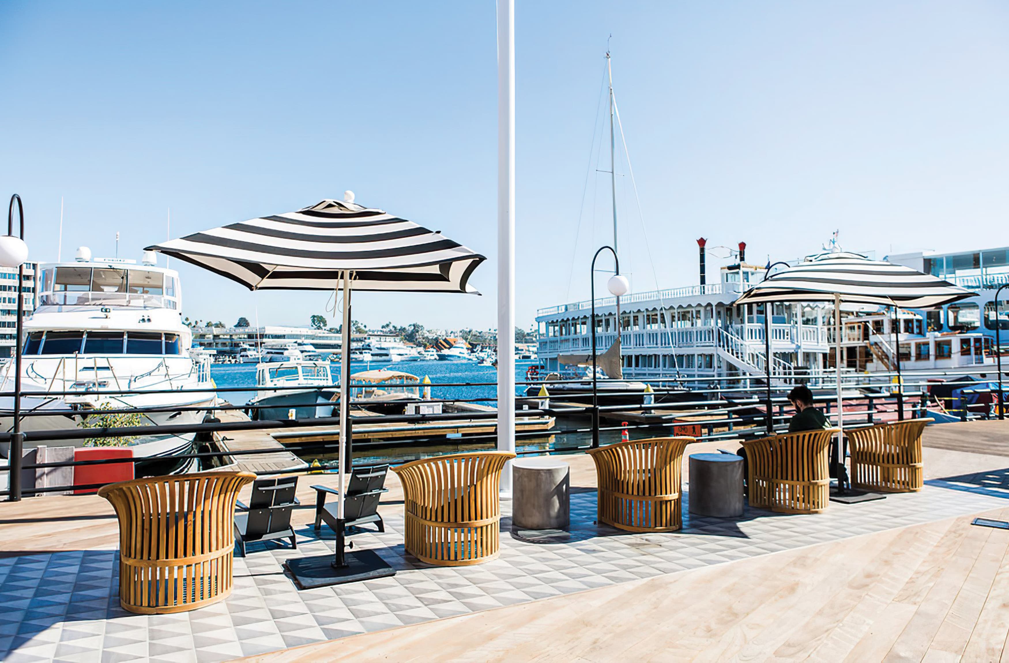 A photograph of a harbor scene featuring seating, black and white umbrellas and views of boats.