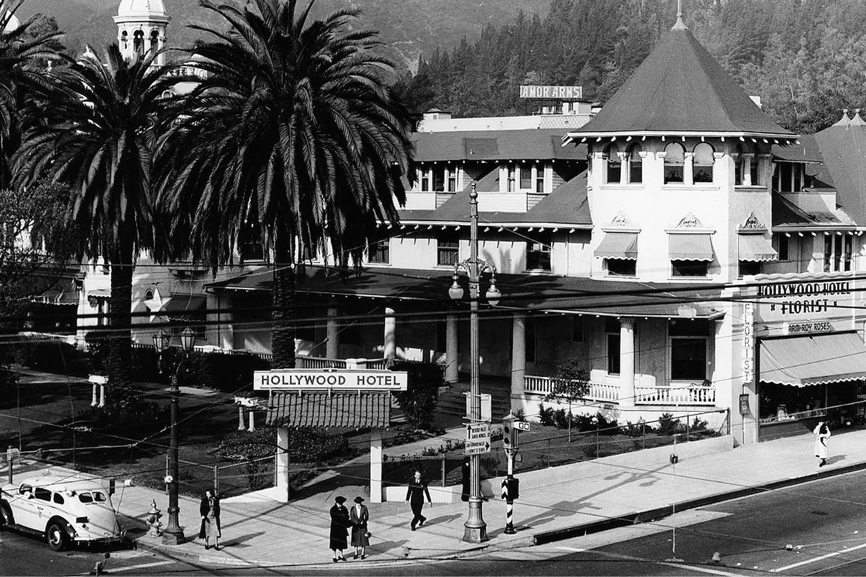 Archival black and white aerial photograph of the historic Hollywood Hotel, a grand Victorian-era landmark at the corner of Hollywood Boulevard and Highland Avenue, featuring a distinctive conical turret roof, wraparound verandas, striped awnings, Hollywood Hotel Florist signage, Amor Arms building in background, tall California fan palms, period 1930s automobiles, and elegantly dressed pedestrians on the sidewalk, capturing early Hollywood California history