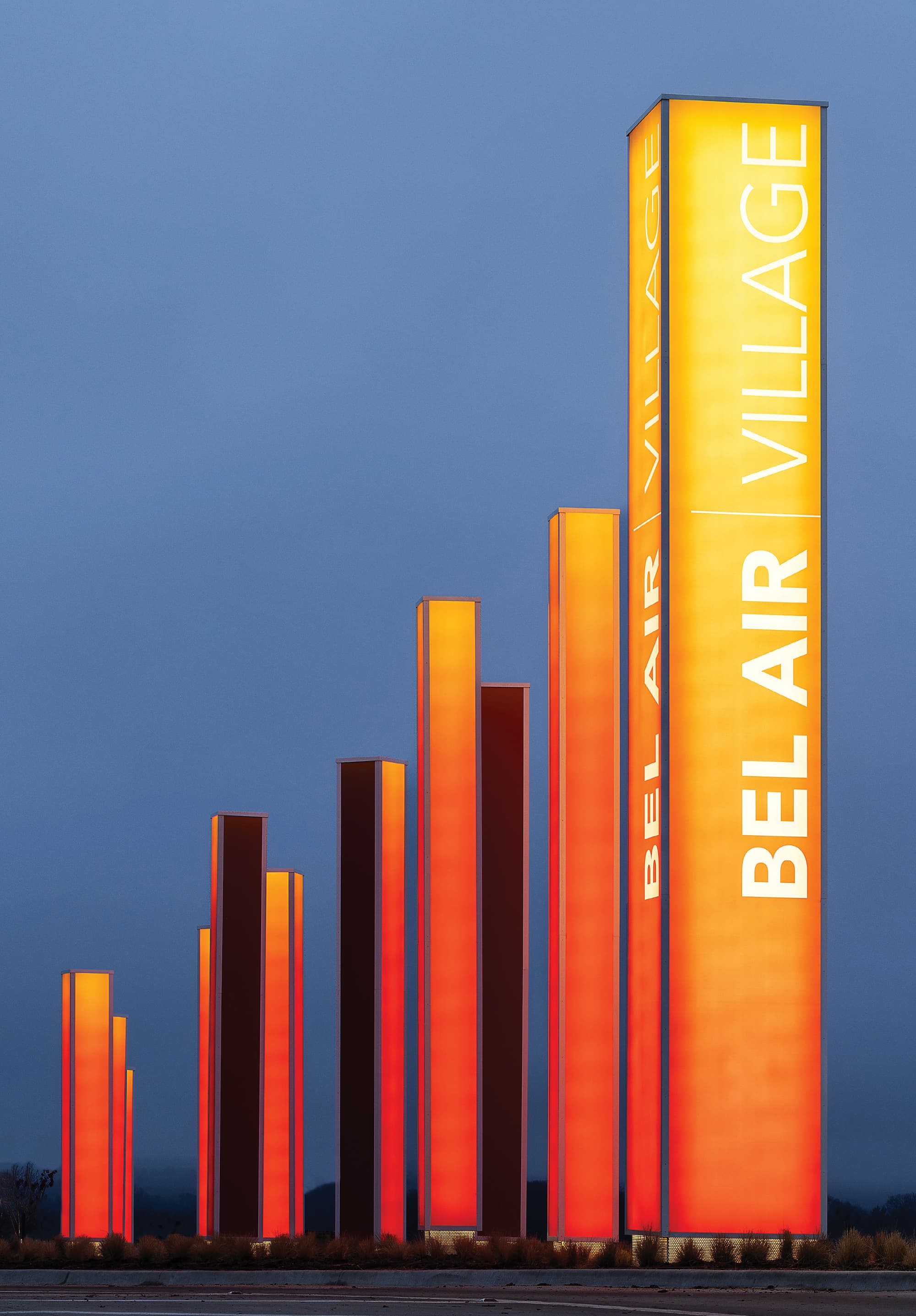 A row of nearly a dozen pylons trail off into the distance at dusk. The nearest, rightmost pylon has text reading “Bel Air Village”, rotated 270-degrees and running vertically from bottom to top. All pylons are illuminated with an yellow-to-orange gradient.