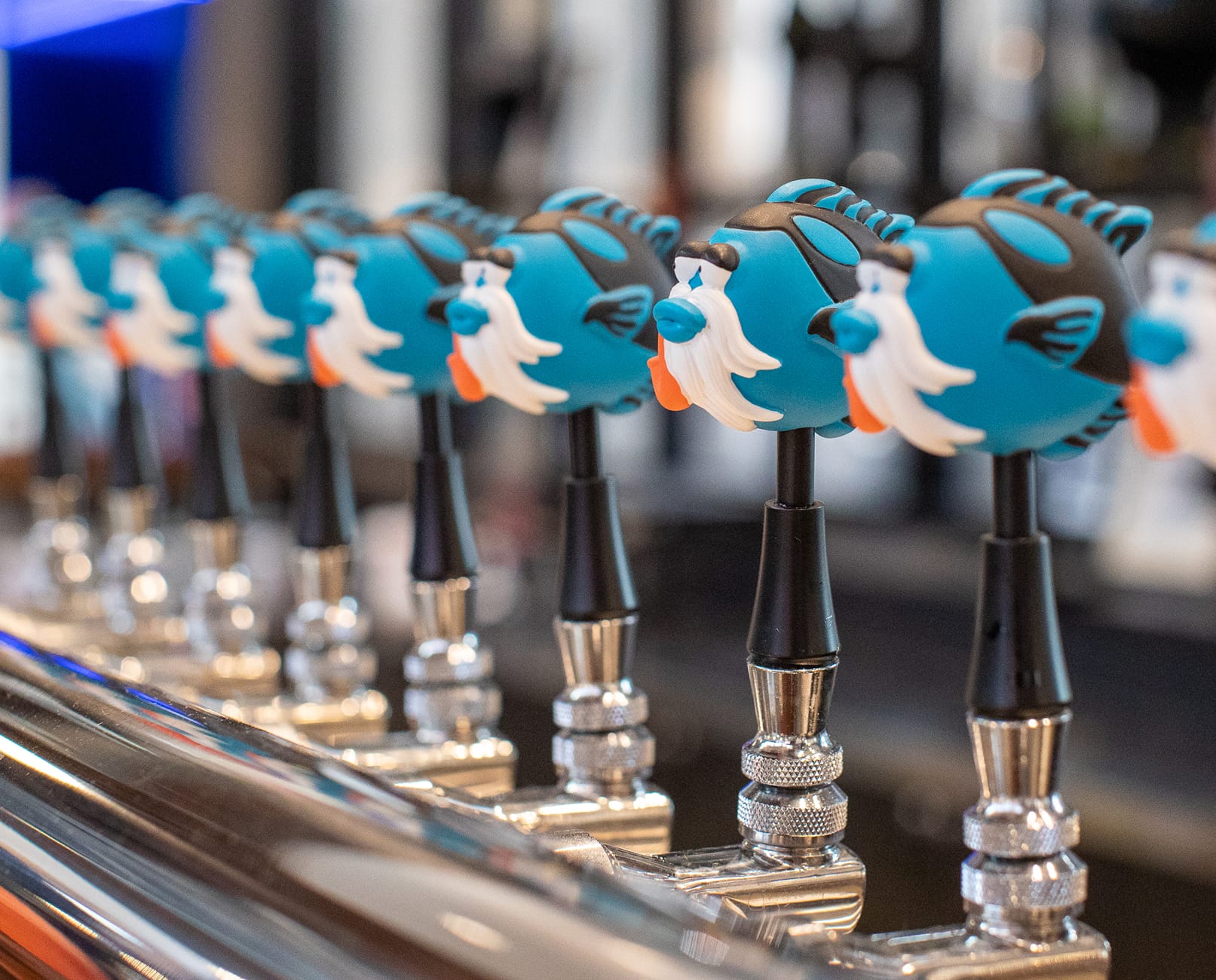 Close-up of a row of custom Bearded Tang Brewing draft tap handles, each shaped as the brewery's bearded tang fish mascot in teal, black, and white, mounted on a polished chrome tap rail at Rodeo 39 Public Market.