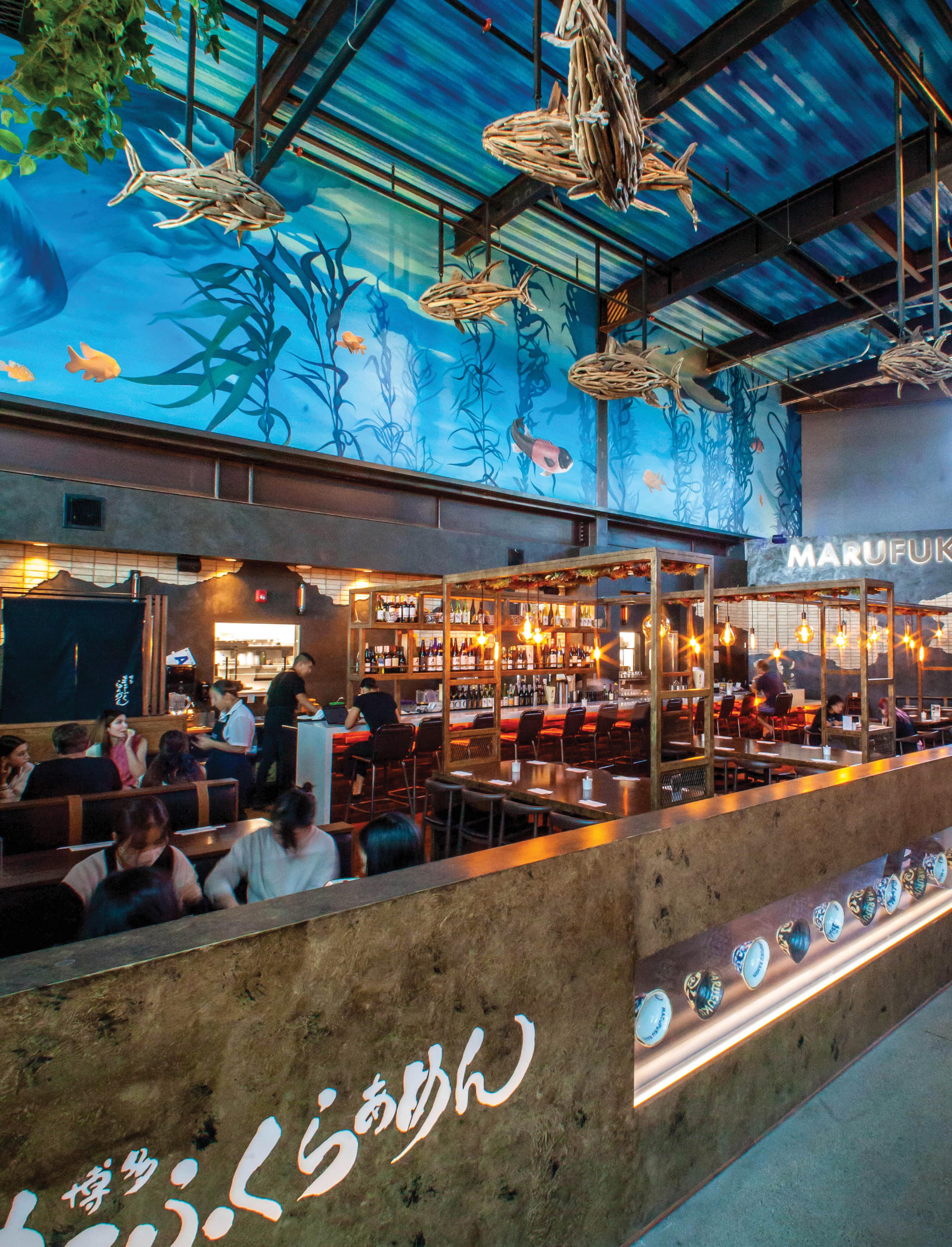 Interior of Marufuku Ramen restaurant featuring an expansive underwater-themed mural with kelp, fish, and ocean imagery spanning the ceiling and upper walls, suspended driftwood fish sculptures, Japanese typography applied to a textured bar front, and warm Edison bulb lighting over a fully seated dining room — demonstrating RSM Design's experiential graphics and branded environments, placemaking, and art programs and public art services within the food and beverage and mixed-use markets