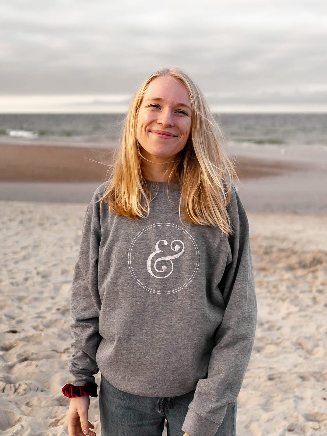 Person wearing a branded gray crewneck sweatshirt featuring the 2nd & PCH ampersand medallion mark in white, photographed on a beach — an example of RSM Design's print and digital collateral and branding and visual identity extending into lifestyle merchandise for a coastal waterfront retail and mixed-use destination in Long Beach, California.