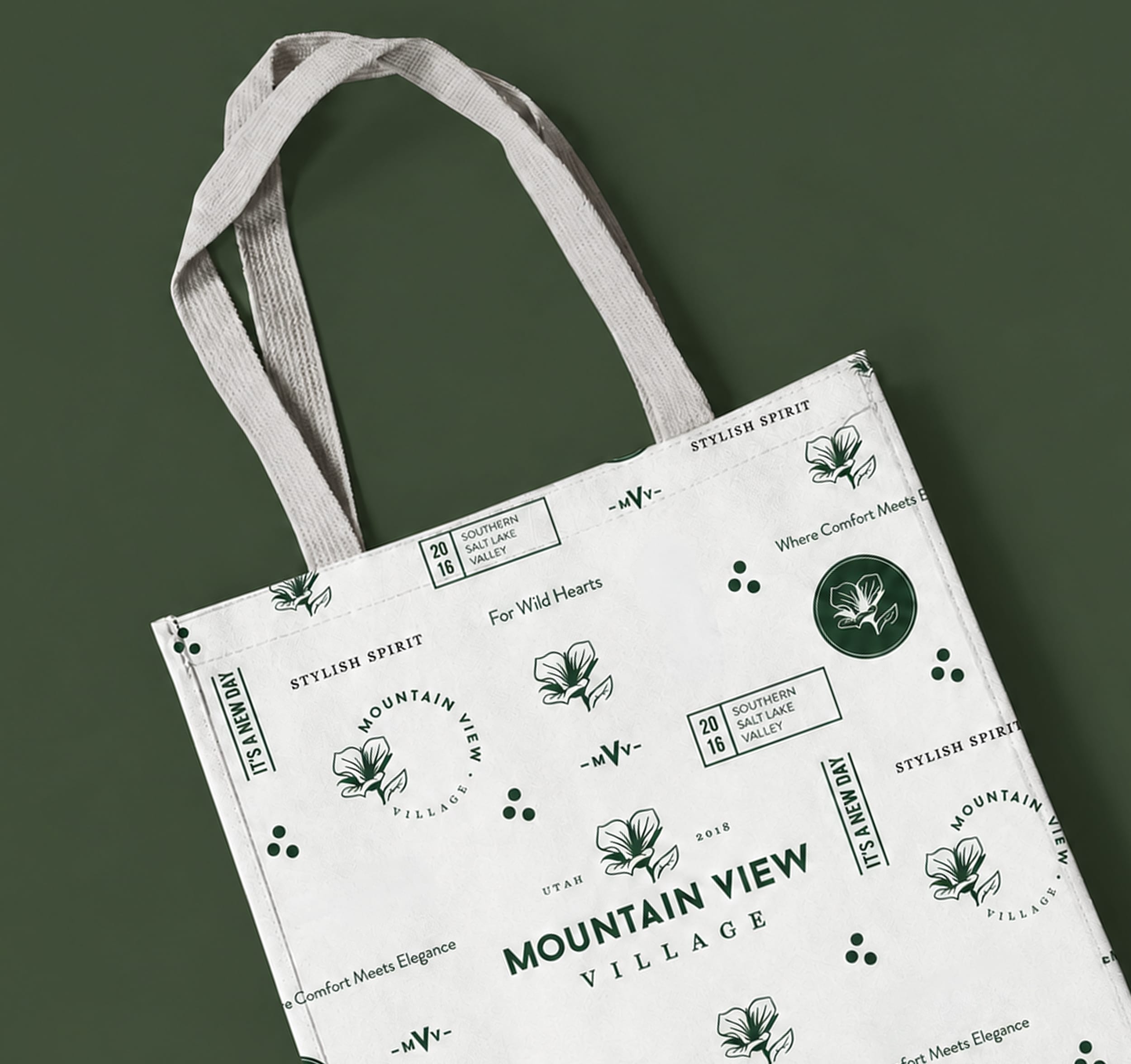 Branded canvas tote bag featuring the Mountain View Village wordmark, sego lily emblem, and pattern of taglines and location details in green and cream — branding and environmental graphic design by RSM Design.