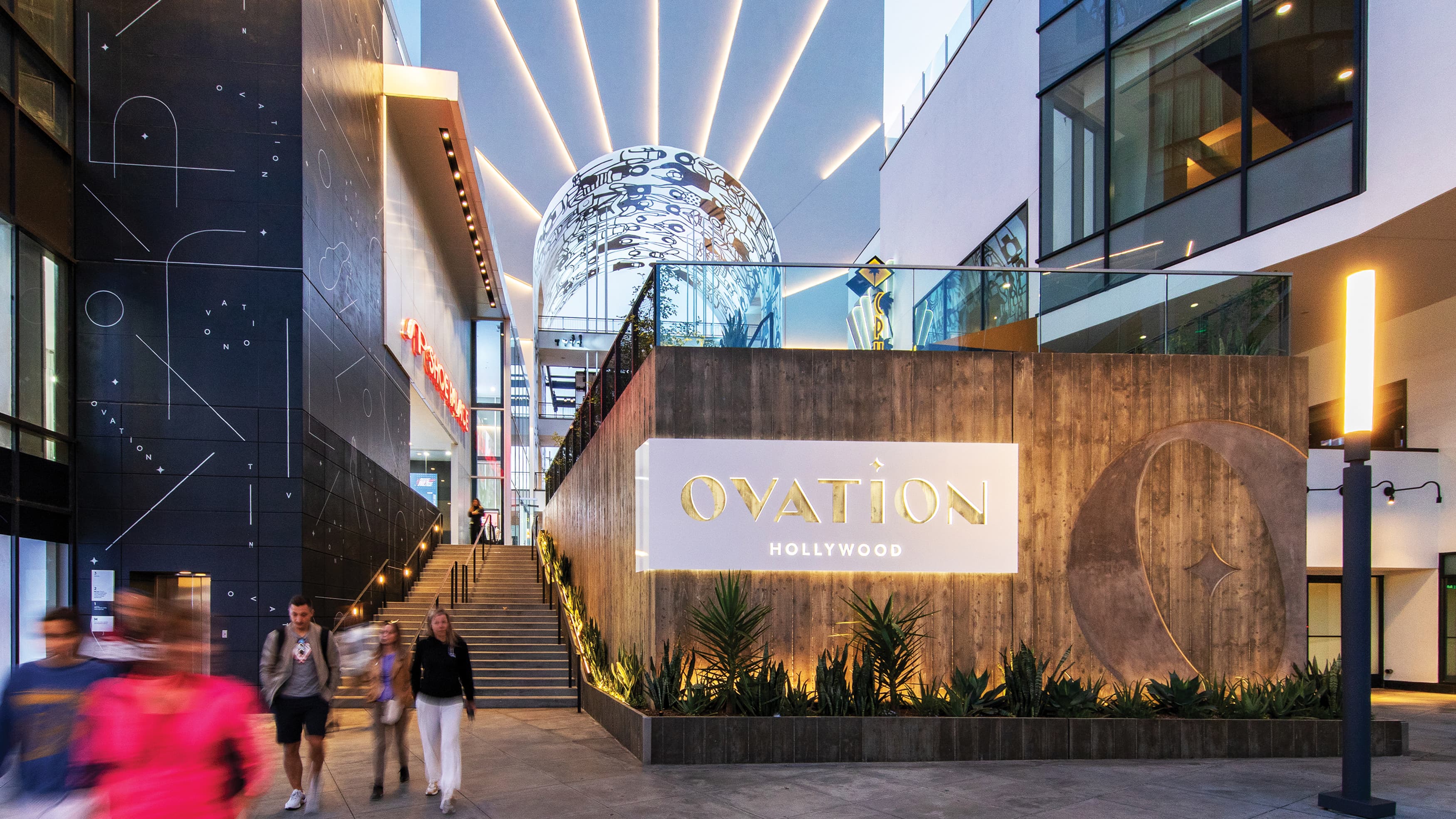 White illuminated Ovation logo sign on board form concrete wall with arch in background with illuminated rays extending on arch face.