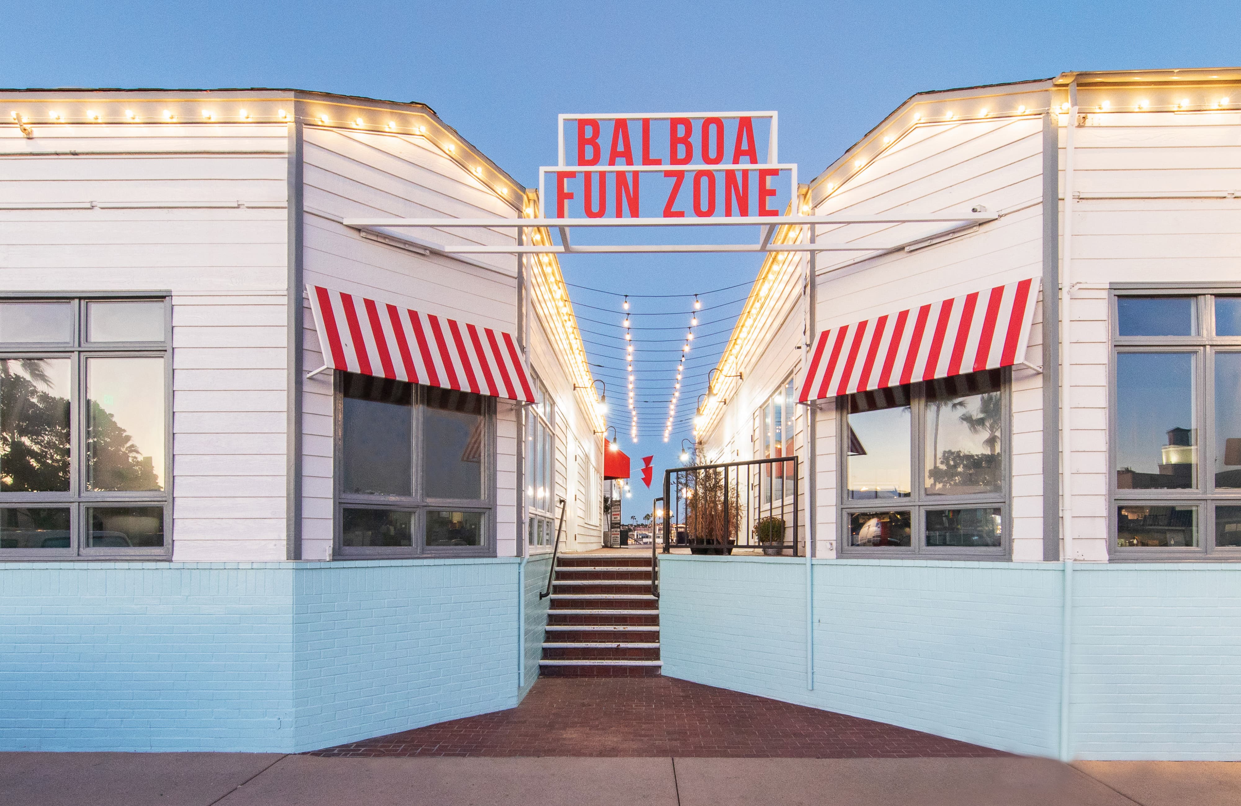 The entry gateway to Balboa Fun Zone at dusk featuring a bold red illuminated "Balboa Fun Zone" identity sign spanning a pedestrian paseo between two white-painted buildings with red and white striped awnings, string lights overhead, and brick steps leading toward the waterfront — showcasing RSM Design's identity signage, branding and visual identity, and placemaking services applied within the waterfront, retail, and mixed-use markets