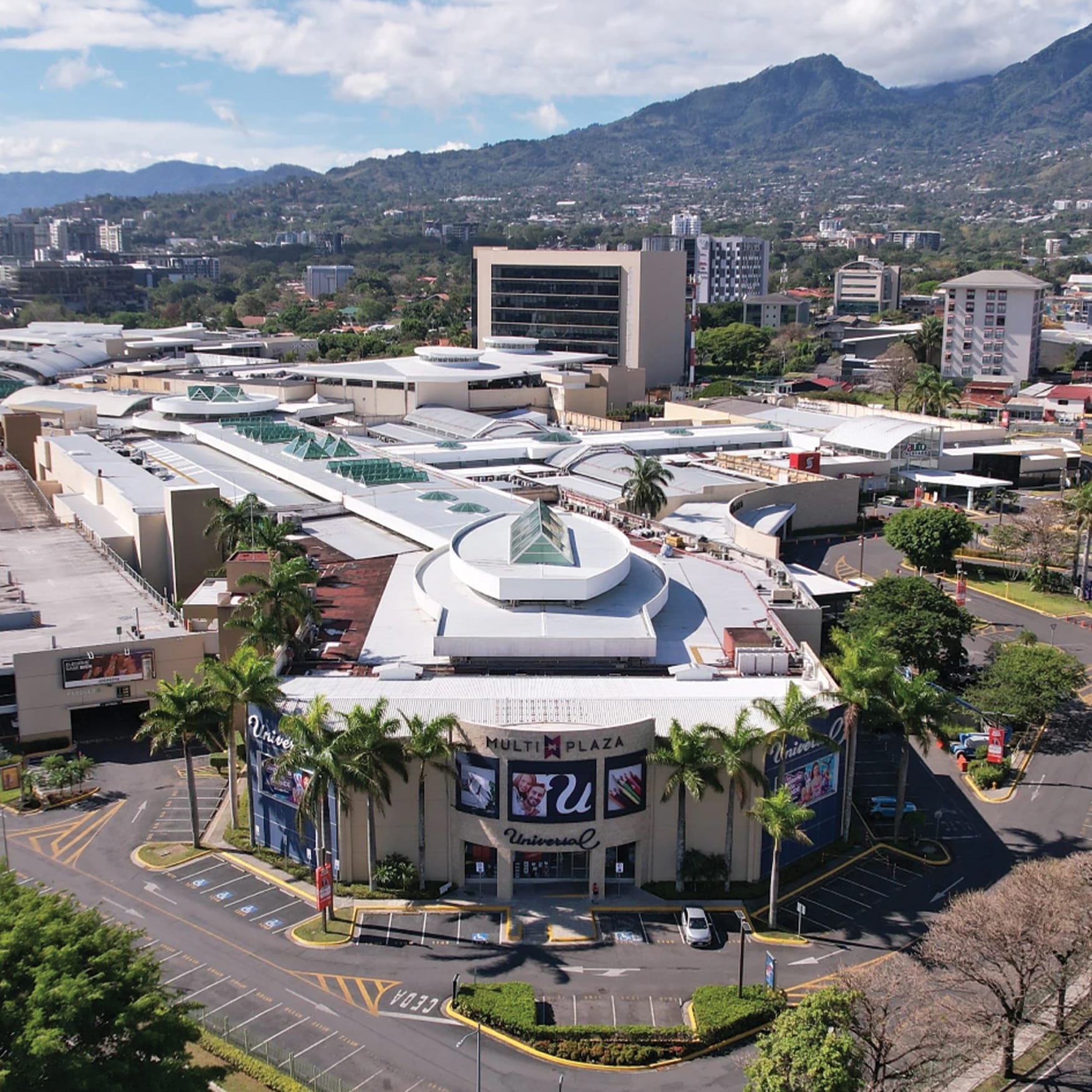 Aerial view of Multiplaza shopping center in Honduras featuring a modern retail complex with curved architecture, skylights, palm-lined entrances, landscaped parking areas, and a mountainous tropical city backdrop.