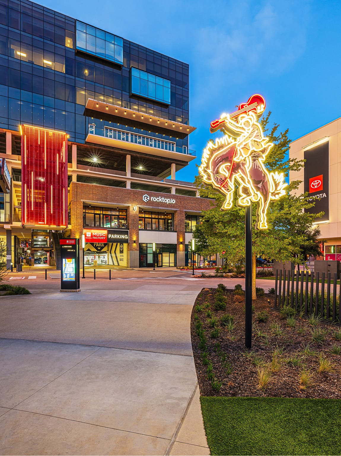 Illuminated neon cowboy guitarist landmark sculpture, digital wayfinding kiosk, and branded parking signage at Toyota Music Factory mixed-use entertainment district in Irving, Texas at dusk — RSM Design environmental graphic design, wayfinding and signage design, specialty graphics, and experiential placemaking for mixed-use, parking garage, and streetscape destinations.