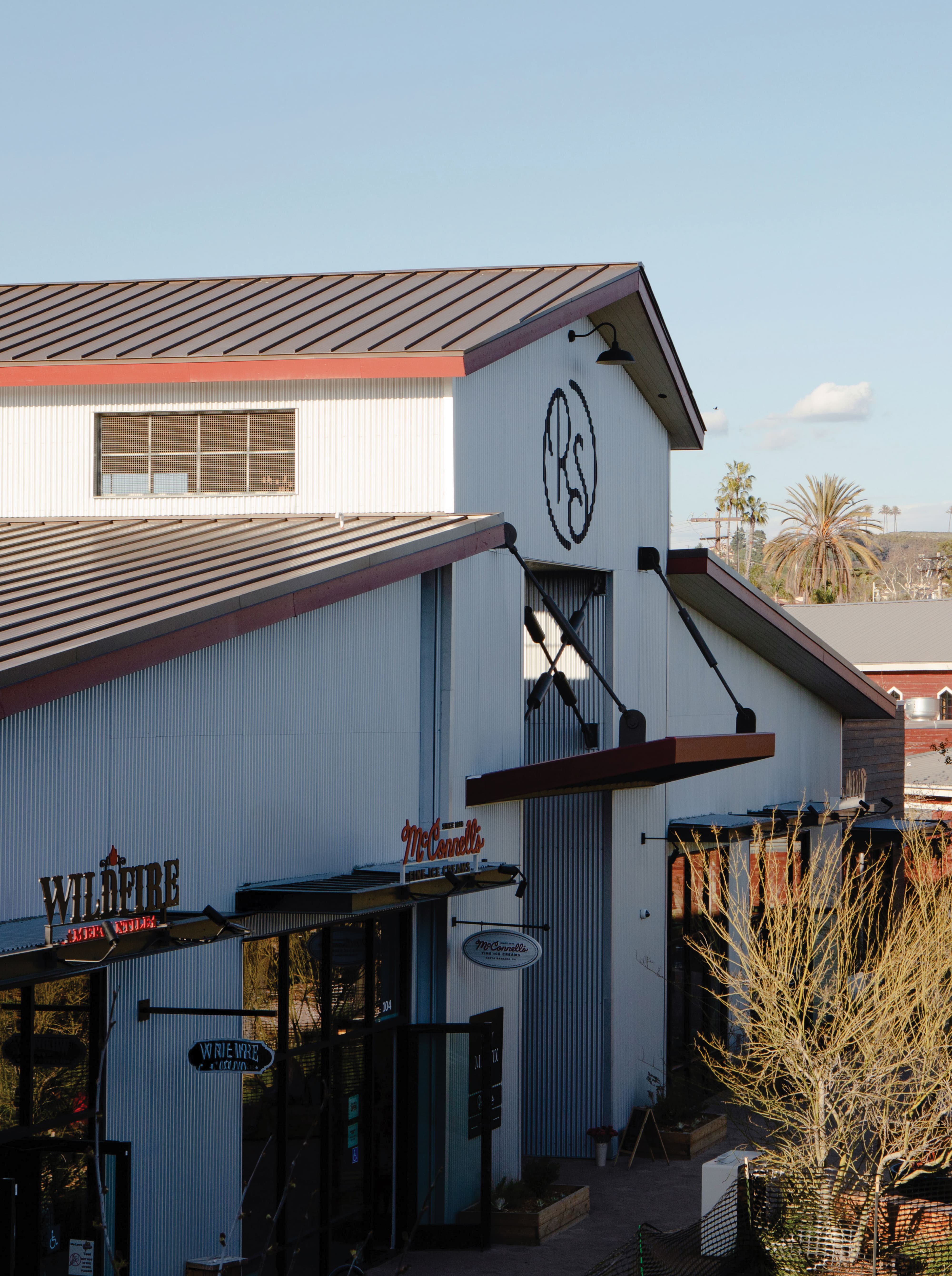 RSM Design environmental graphic design and master sign program for River Street Marketplace, featuring an exterior view of a multi-tenant mixed-use building with a large-scale crossed oar placemaking installation and RS monogram logo, alongside tenant blade signs for Wildfire and McConnell's Fine Ice Cream, integrating wayfinding signage, experiential graphics, and branded environments into a cohesive food and beverage and retail streetscape.