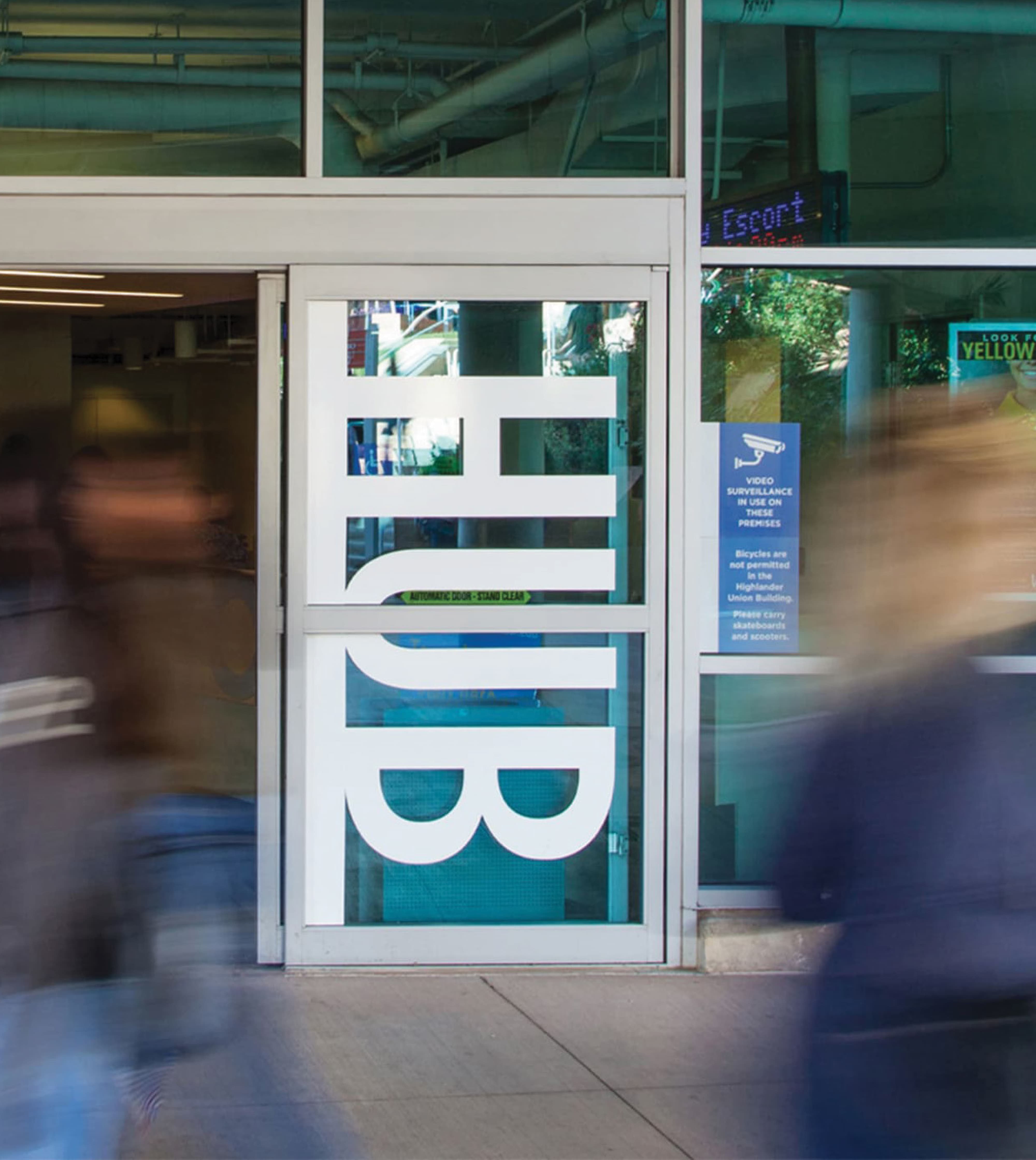Automatic glass door with 'HUB' lettering and building policy signage, as blurred figures walk past in the foreground.