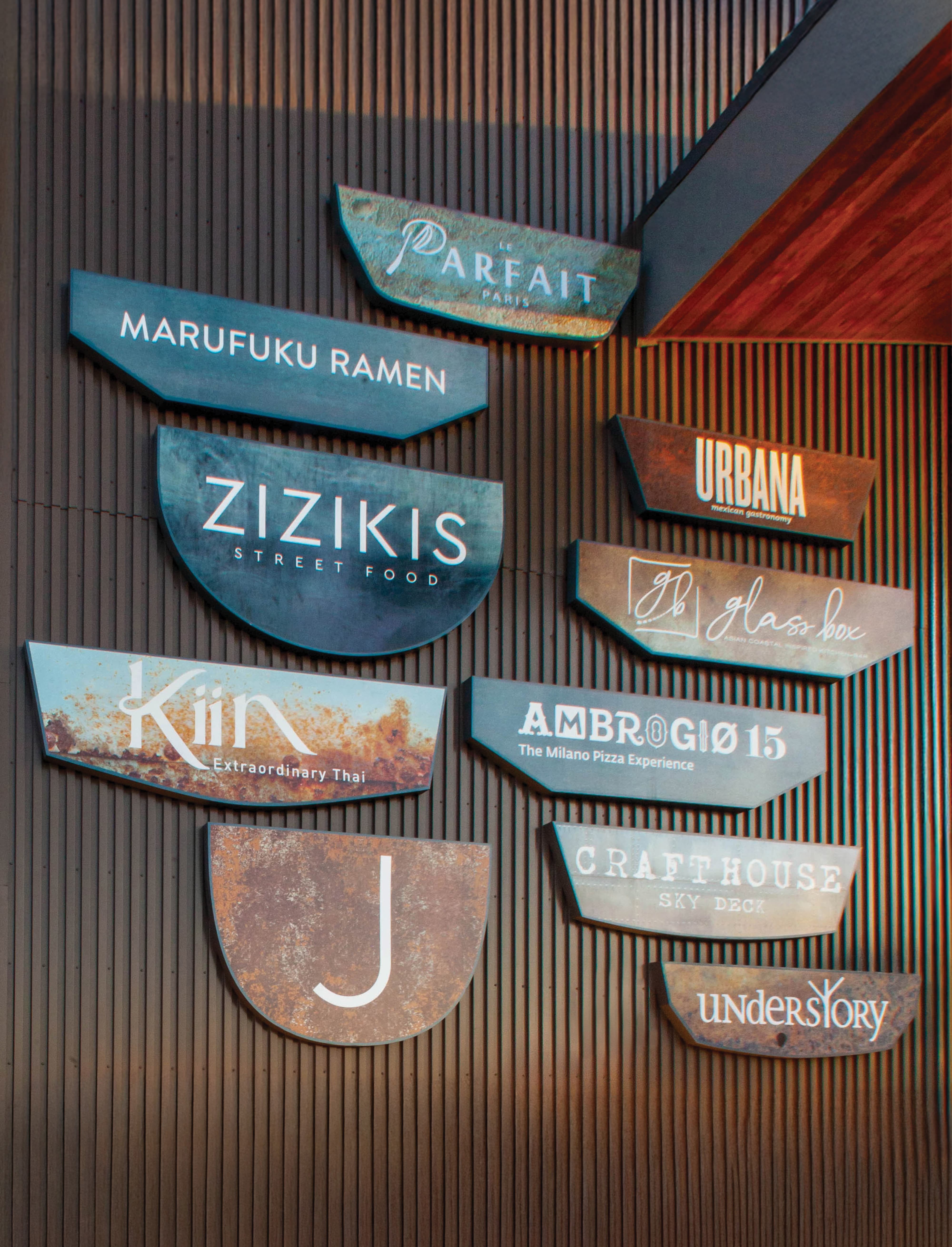 A collection of dimensional tenant identity signs mounted on a corrugated metal wall, featuring restaurant and food and beverage brands including Le Parfait Paris, Marufuku Ramen, Zizikis Street Food, Kiin Extraordinary Thai, Urbana Mexican Gastronomy, Glass Box, Ambrogio15, Crafthouse Sky Deck, Understory, and others — each rendered in unique materials, patinas, and typographic styles — showcasing RSM Design's tenant signage, master sign programs, and experiential graphics and branded environments across food and beverage and mixed-use markets
