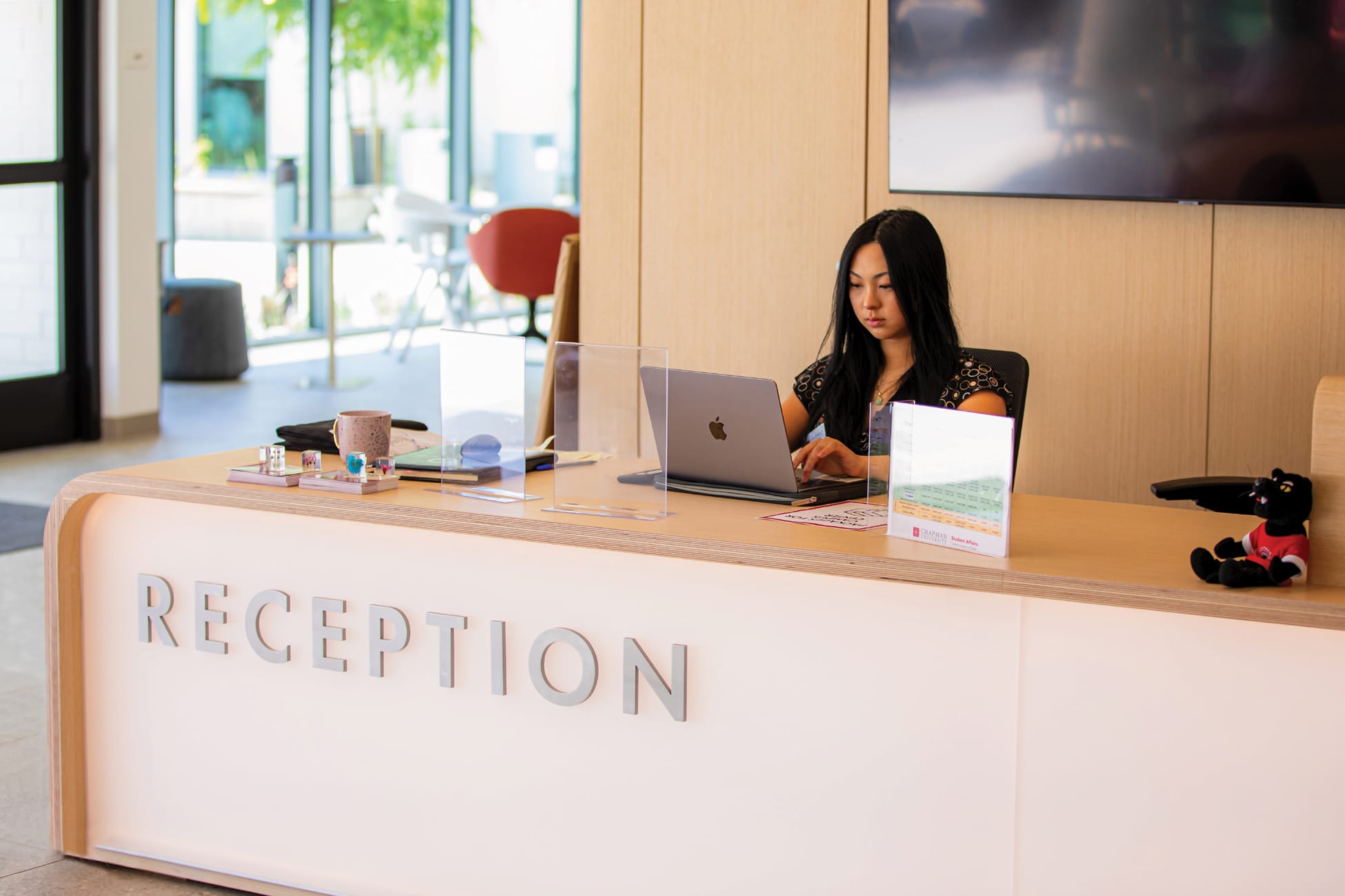 RSM Design Chapman University Rinker Campus Center Irvine — student staffed reception desk with dimensional brushed metal letter signage, warm plywood fascia, and open campus views beyond