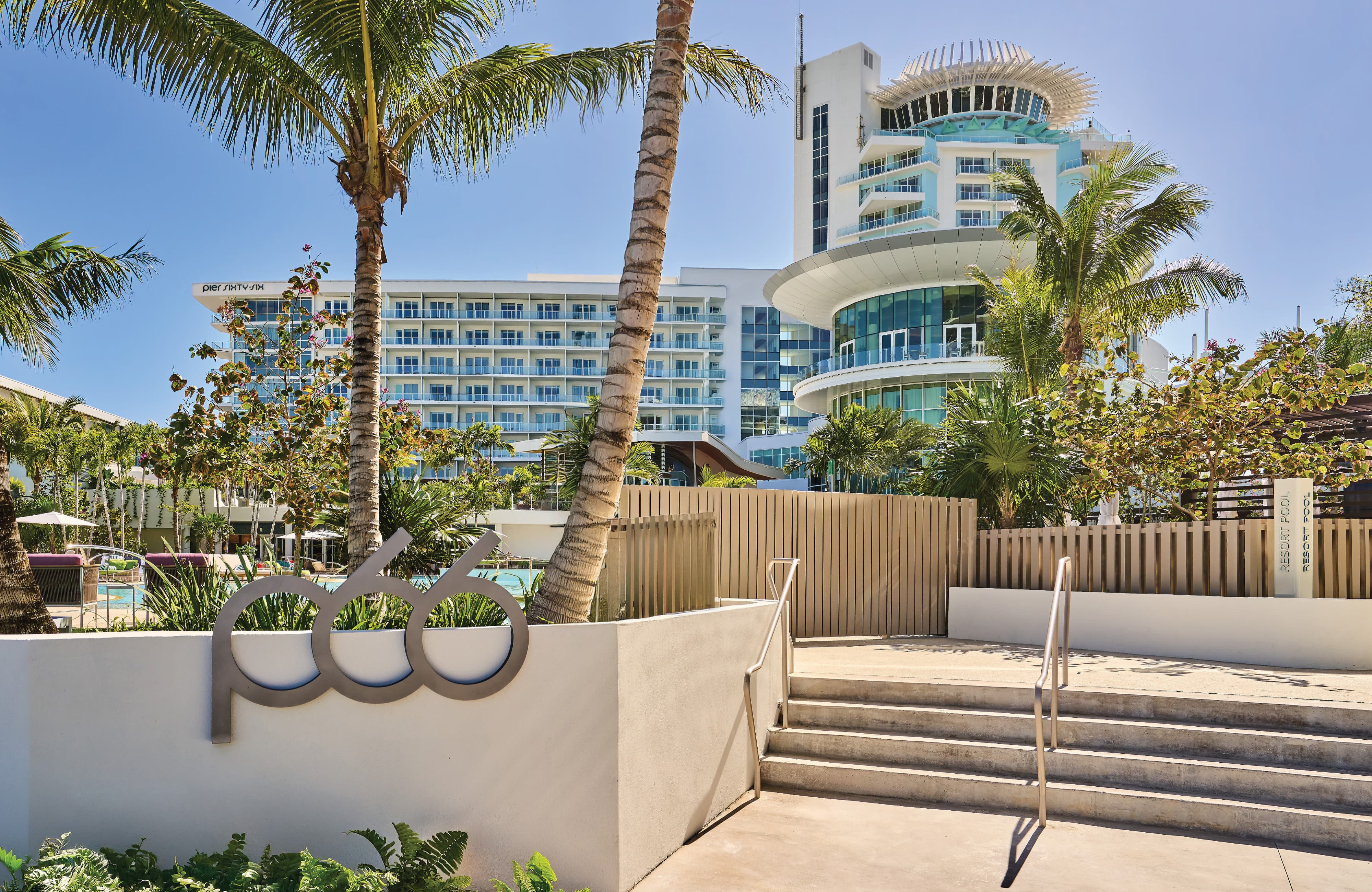 Dimensional metal "p66" logo sign mounted on a poolside planter wall at Pier Sixty-Six luxury waterfront resort in Fort Lauderdale, Florida, with iconic rotating tower and tropical landscaping in background — RSM Design branding and visual identity, wayfinding and signage design, specialty graphics, and experiential placemaking for hospitality and waterfront destinations.