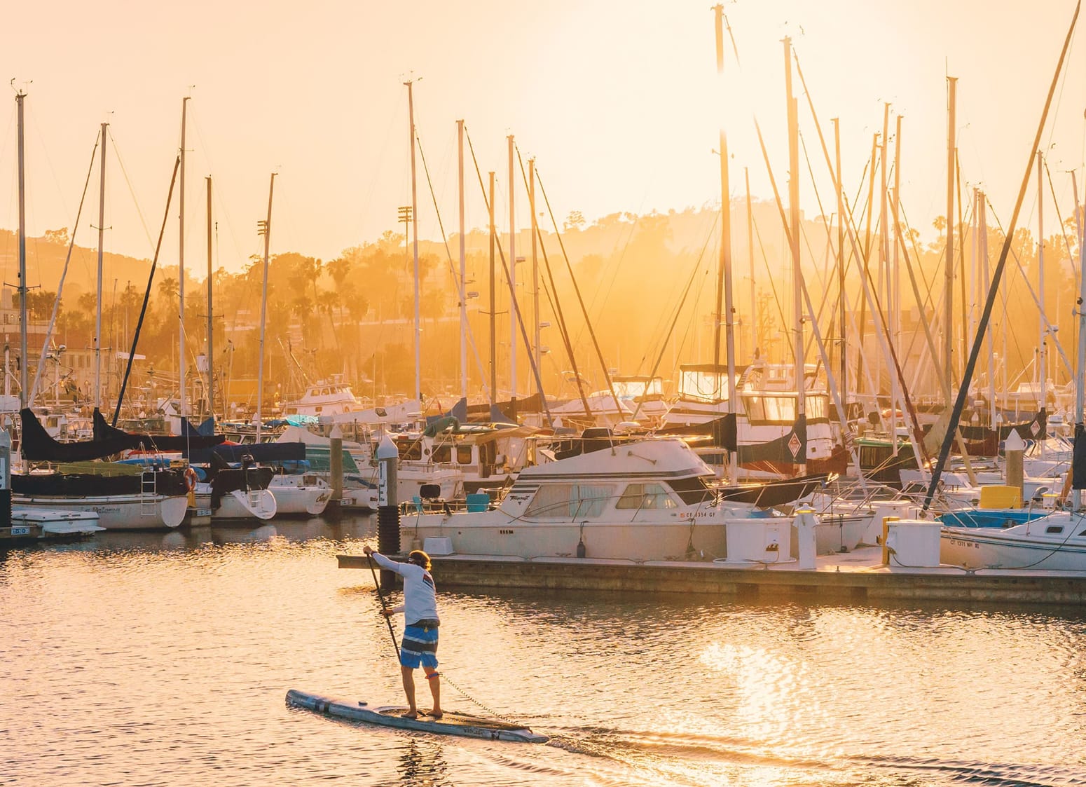 Paddleboarder crossing the Dana Point Harbor marina at sunset with sailboats lining the docks