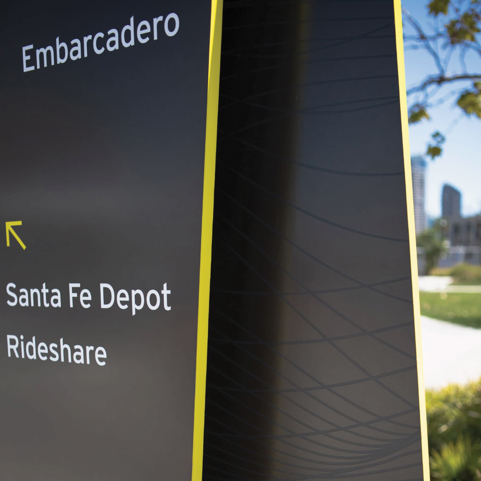 Close-up of a black wayfinding sign with yellow edge accents, reading 'Embarcadero' and an arrow pointing to 'Santa Fe Depot' and 'Rideshare,' with a park and city skyline visible in the background.