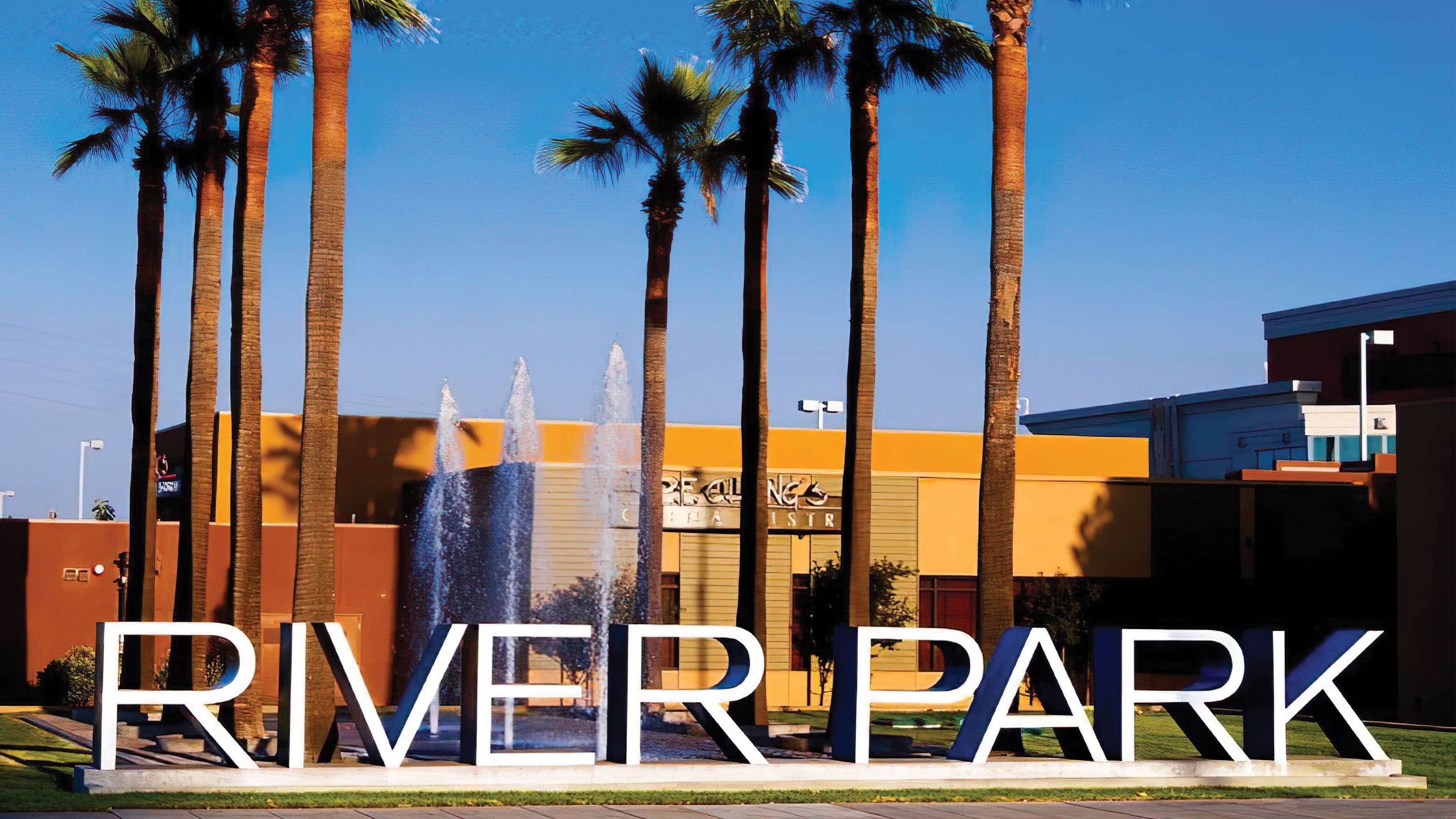 Large, dimensional Riverpark placemaking identity with white face and blue return flanked by a fountain and palm trees