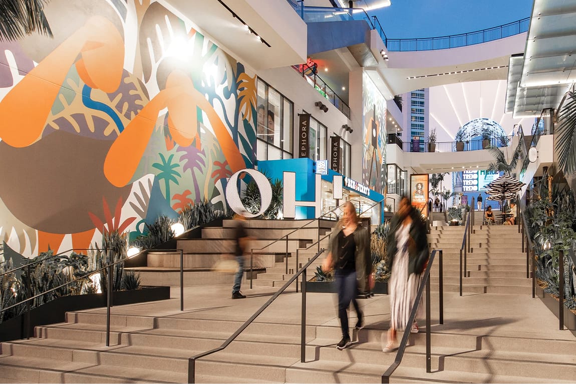 Twilight interior view of Ovation Hollywood's multilevel open-air retail terrace staircase featuring a large-scale vibrant tropical figurative mural with bold orange, teal, and green botanical motifs, illuminated three-dimensional "OH" letter sculpture installation, Sephora store signage, Dick's Sporting Goods, native succulent and agave landscaping in stepped planters, motion-blurred shoppers ascending concrete stairs with black steel railings, the iconic LED globe visible in the background, and striped market umbrellas in the upper dining terrace, Hollywood Boulevard Los Angeles