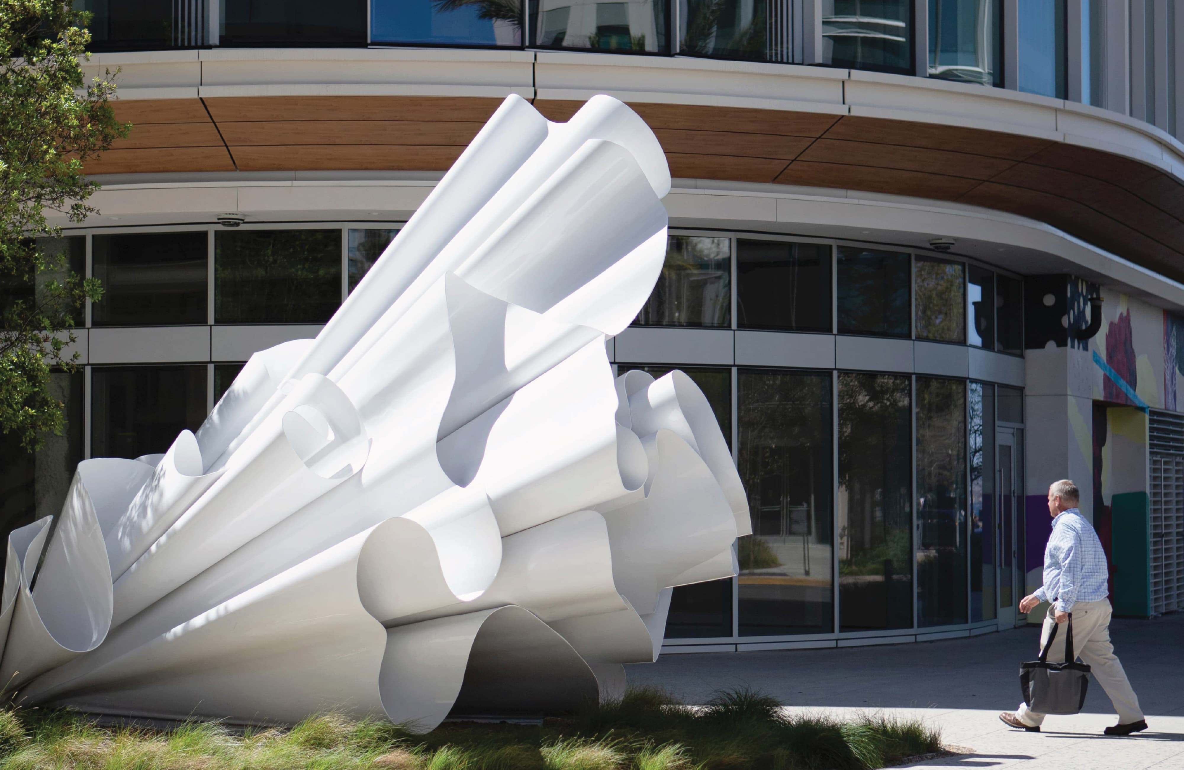 Large sculptural white artwork with sweeping, ribbon-like folds resembling a burst of fabric, installed outside a curved modern building as a man walks past carrying a tote bag.