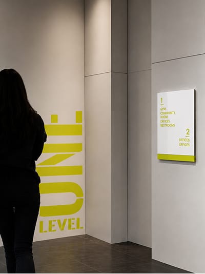 Interior lobby with a large yellow 'Level One' graphic on the wall, a silhouetted person walking past, and a white directory sign listing Level 1: Gym, Community Room, Offices, Restrooms and Level 2: Offices.