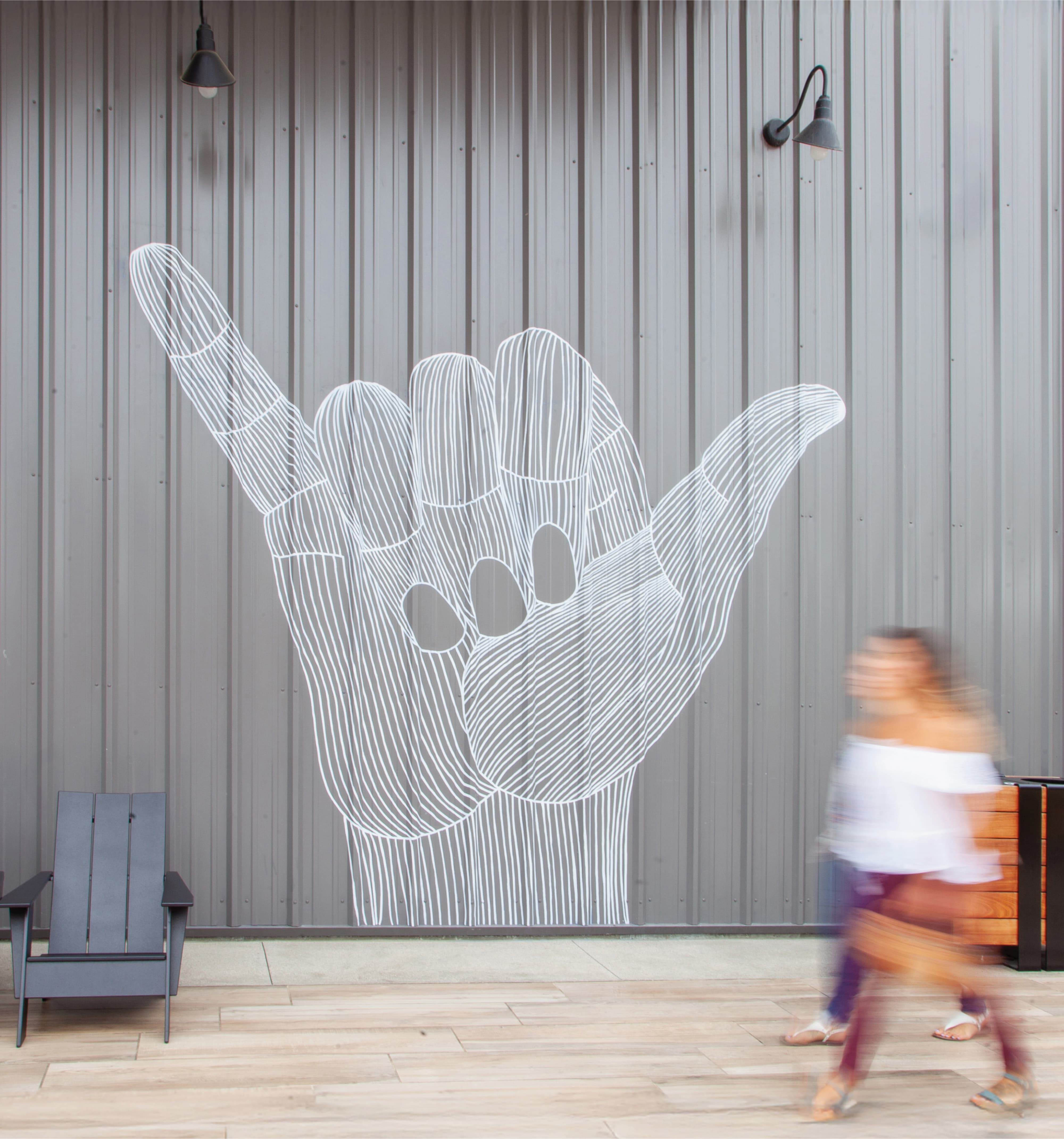 Large line-art mural of a hand making the shaka 'hang loose' sign on a corrugated metal wall, with a black adirondack chair and wall sconces nearby, as a blurred woman walks past in motion.