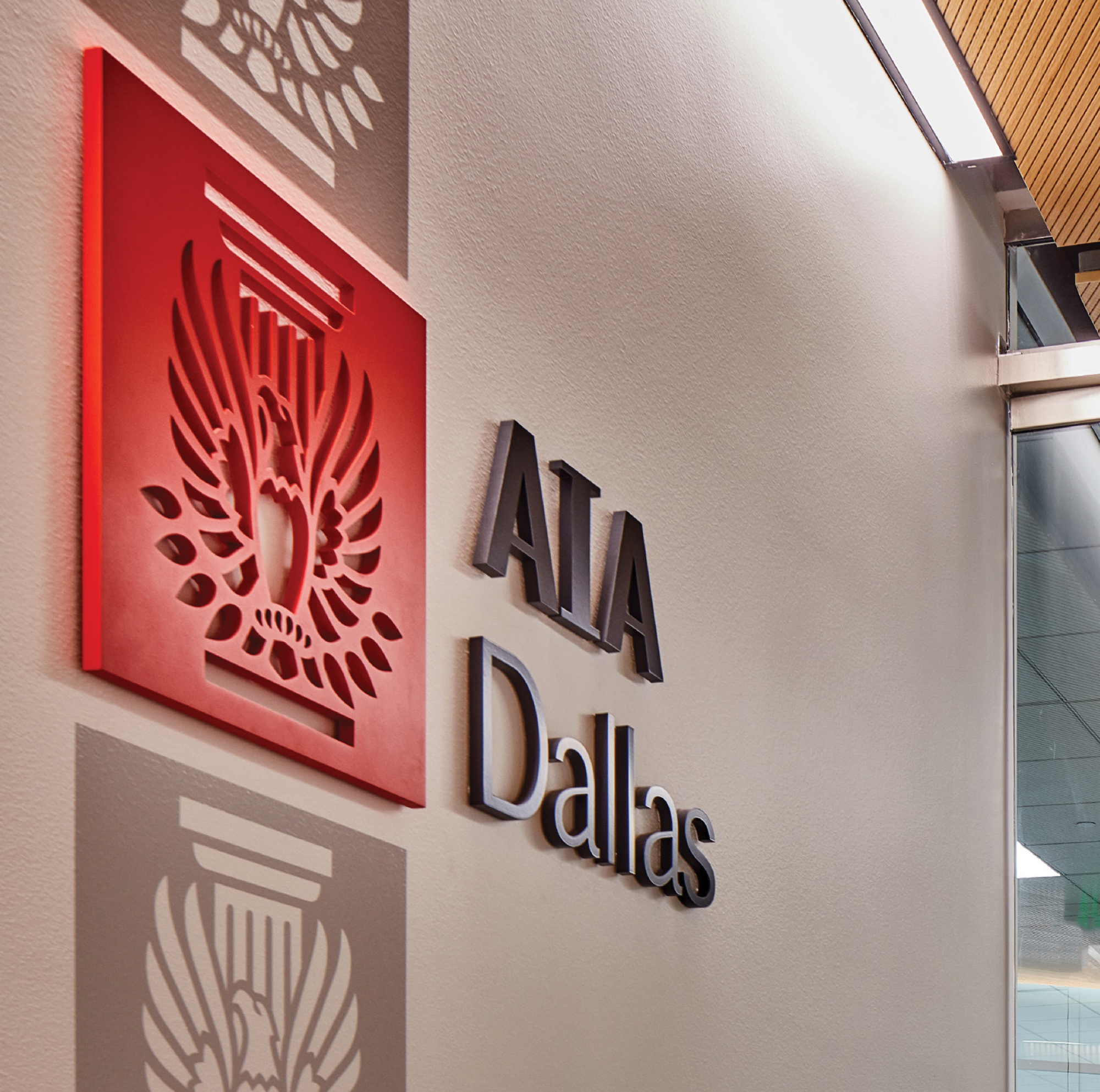 RSM Design interior identity signage and specialty graphics for AIA Dallas at the Architecture and Design Exchange, featuring dimensional dark metal AIA Dallas lettering mounted alongside a layered red dimensional logo relief panel with an intricately routed eagle emblem casting shadow patterns on the wall, with a tonal ghost version of the logo below, demonstrating how environmental graphic design, visual identity, and branded environments bring institutional identity to life through material craft and dimensional depth for cultural, civic, and workplace markets.