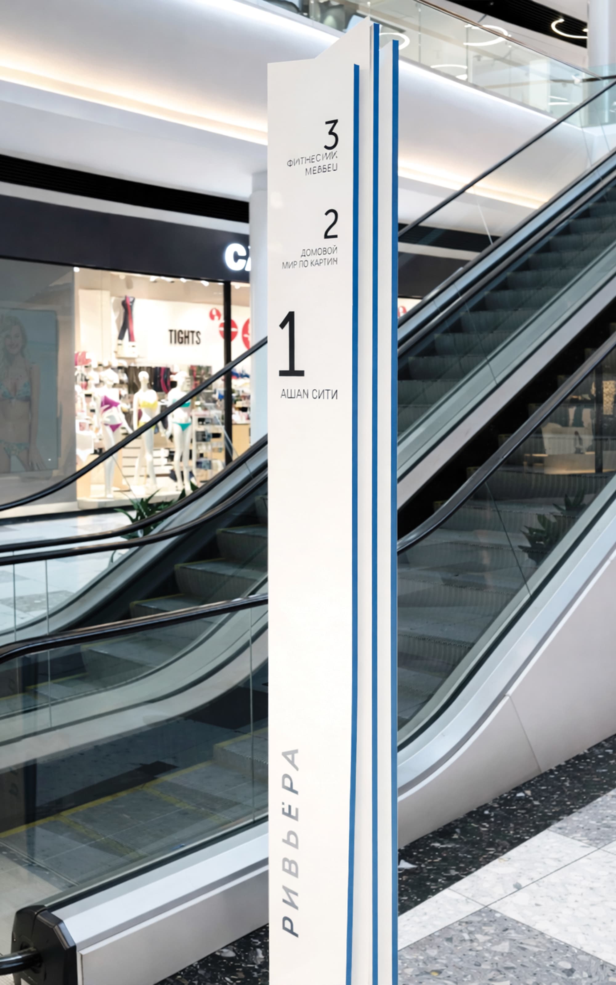 Branded Riviera multi-level floor directory wayfinding pylon sign with Cyrillic level identification adjacent to escalators in an international retail center — RSM Design wayfinding and signage design, master sign programs, environmental graphic design, and branded environments for retail, mixed-use, and international destinations.