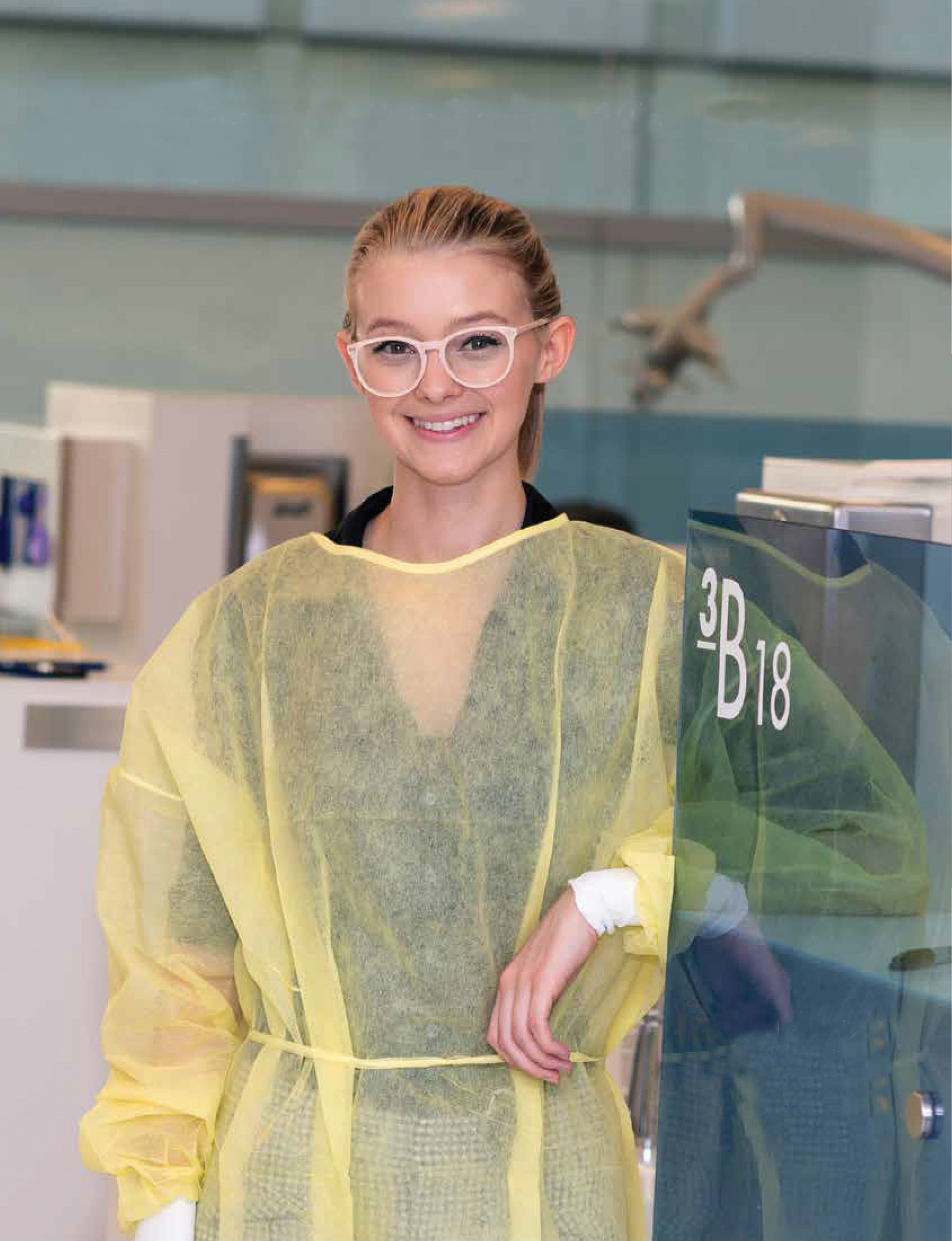 Healthcare employee smiling in a yellow isolation gown beside a wayfinding sign reading "3B 18" in a health and wellness facility, featuring environmental graphic design and signage by RSM Design.