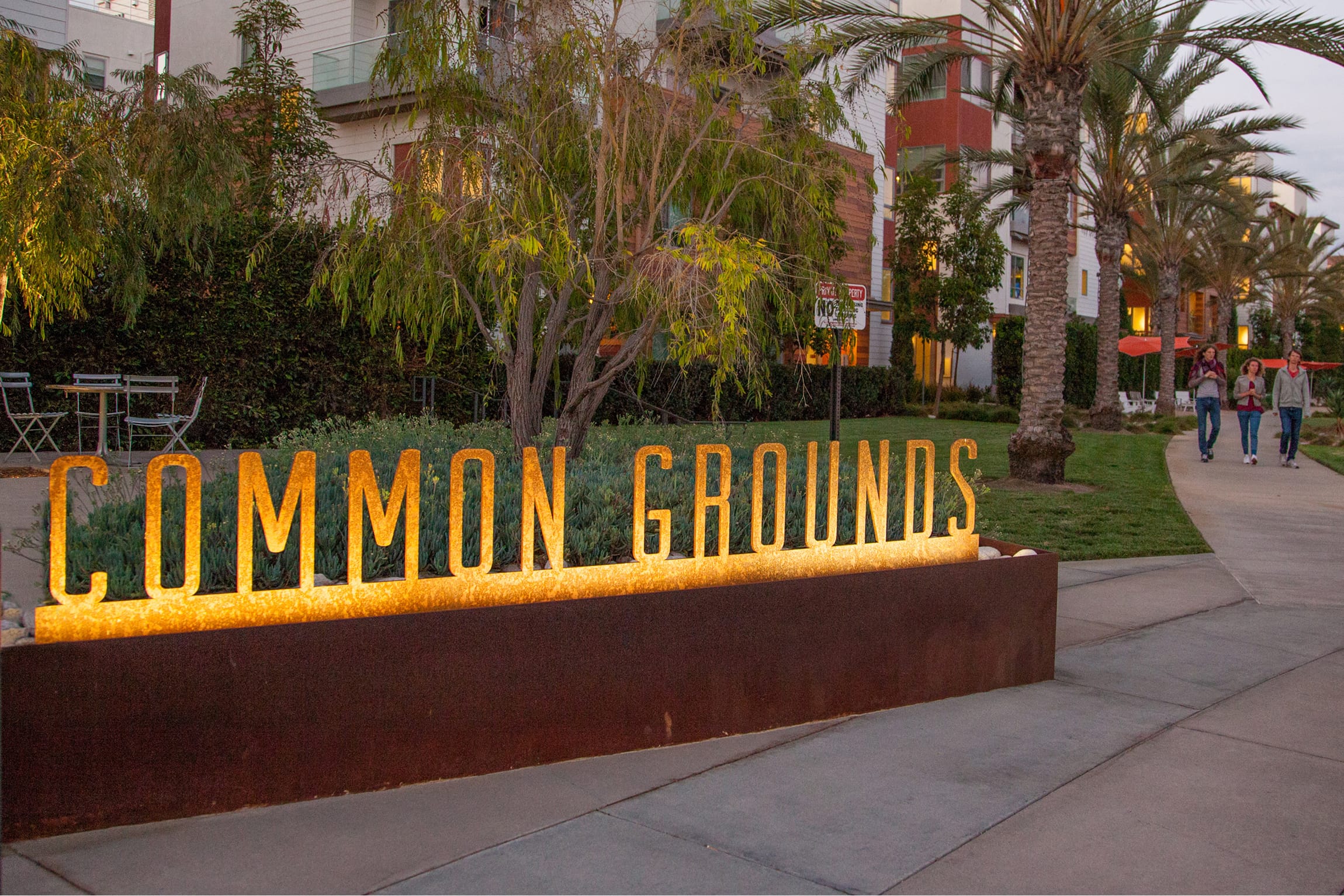 Illuminated corten steel letters spelling "COMMON GROUNDS," designed by RSM Design, set in a planter with palm trees and pedestrians on a walkway in Playa Vista at dusk.