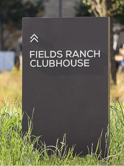 Dark charcoal monolith wayfinding sign with white text and double chevron icon directing guests to Fields Ranch Clubhouse at PGA Frisco
