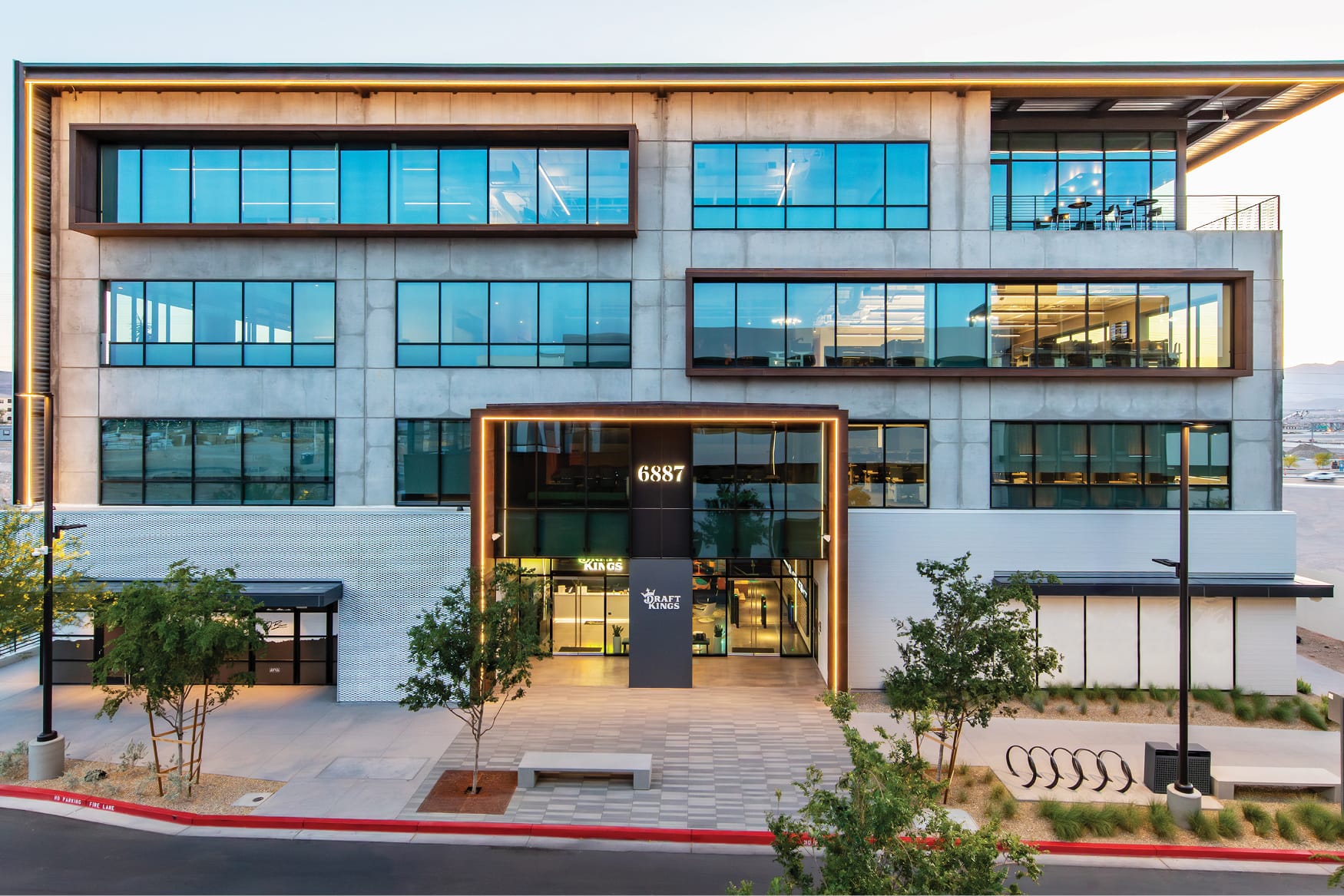 Exterior view of a multi-story office building at 6887 Uncommons in Las Vegas, photographed at dusk. The facade features a concrete and glass curtain wall with Corten steel accents framing large horizontal window bands. A illuminated entrance canopy leads to a DraftKings retail storefront at street level. Young trees, paved plazas, bike racks, and desert landscaping surround the building entrance.

