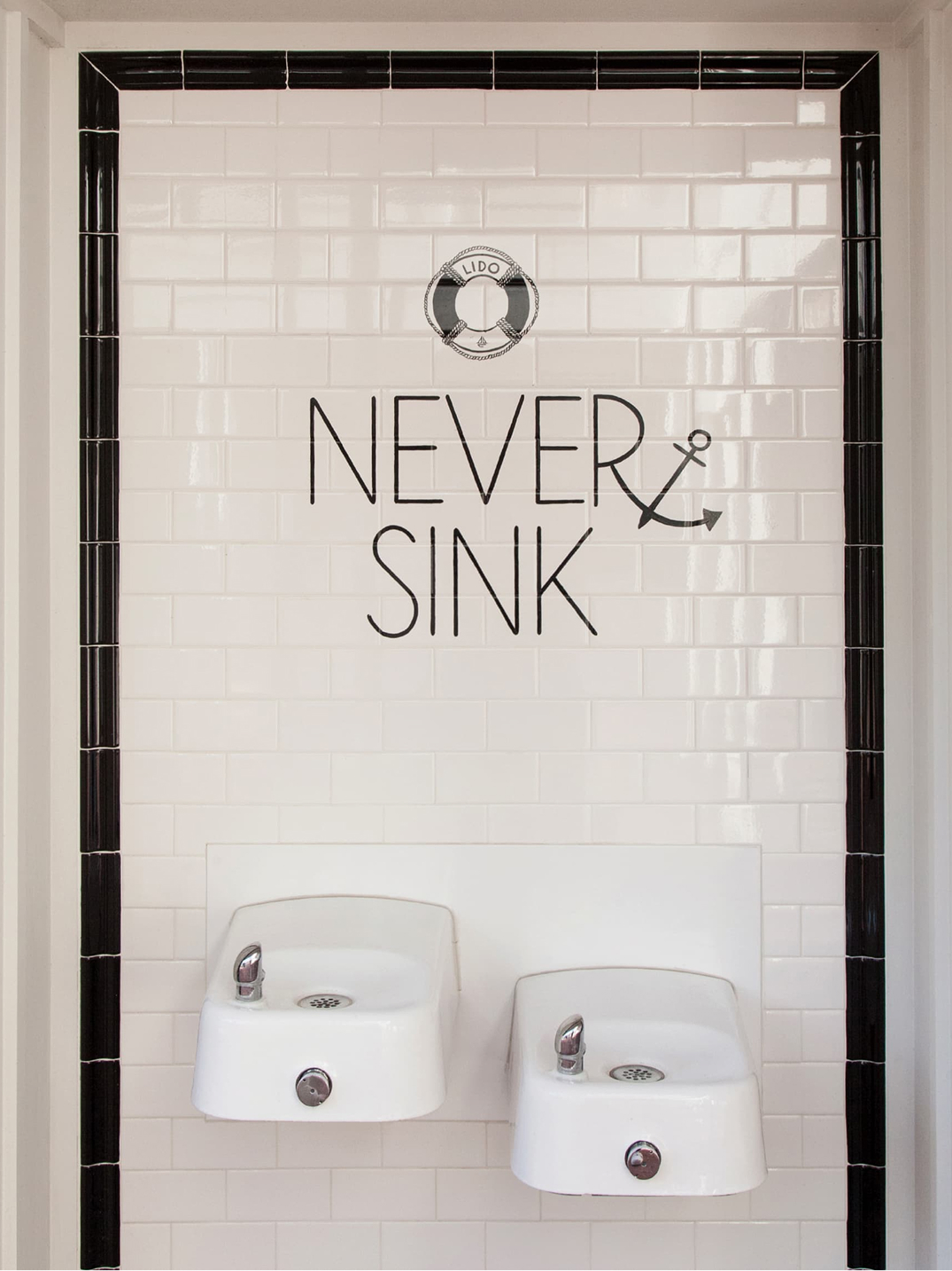 Lido Marina Village public restroom drinking fountains with nautical-themed "Never Sink" typography and life preserver graphic on white subway tile wall with black border, Newport Beach, California